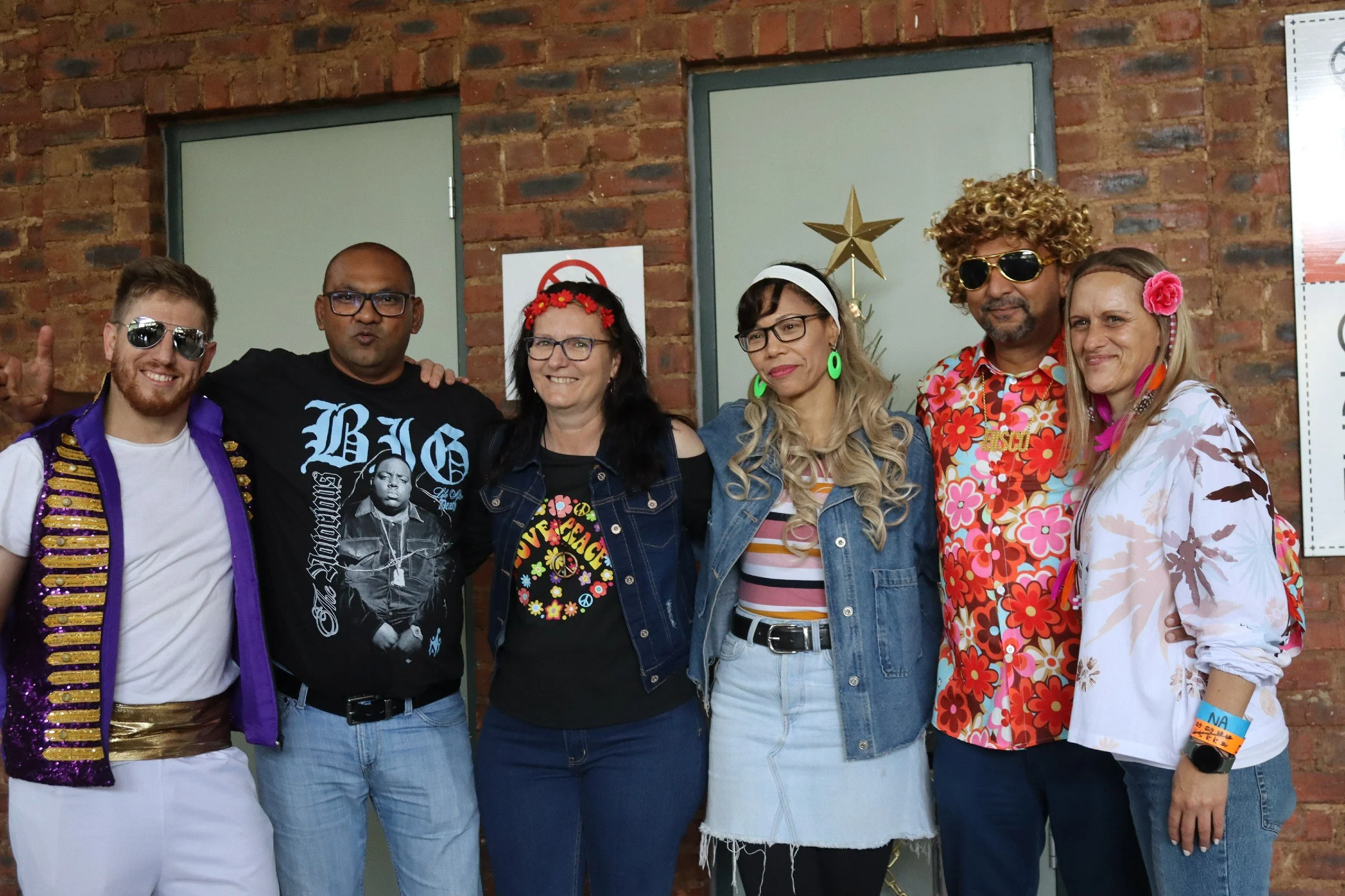 Group of six people dressed in colorful, retro, and floral outfits, standing side by side against a brick wall, smiling and posing for the photo.
