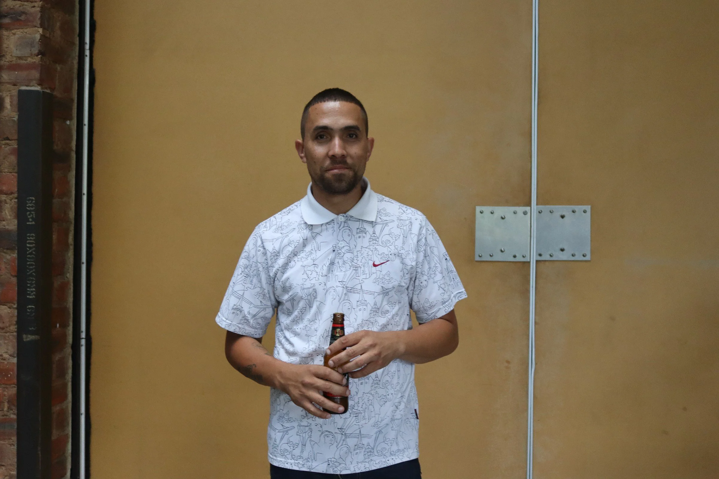 A man with a trimmed beard and short hair, wearing a white T-shirt with a line art design, holding a bottle, standing in front of a yellow wall with a metal panel and exposed brick to the left.