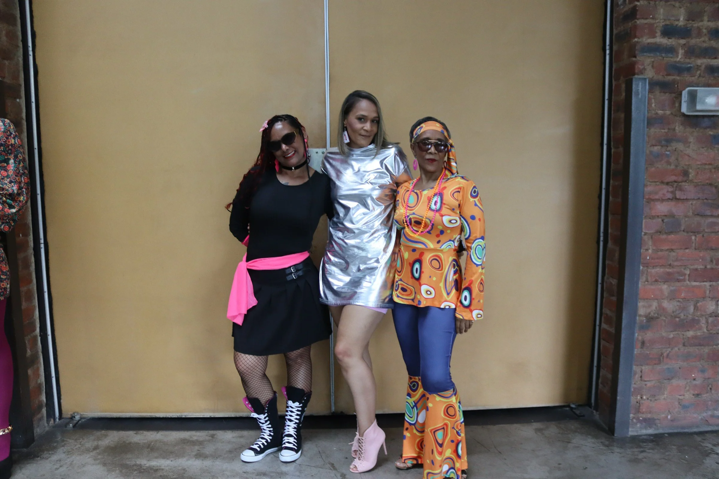 Three women posing in colorful outfits in front of a yellow partition and brick wall.