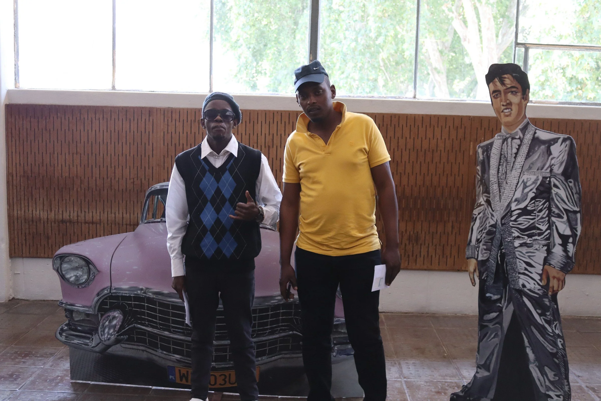 Two men standing indoors in front of a pink vintage car and a cutout of Elvis Presley. The man on the left is wearing a white shirt, a dark vest with an argyle pattern, sunglasses, and a beanie. The man on the right is in a yellow polo shirt with a b