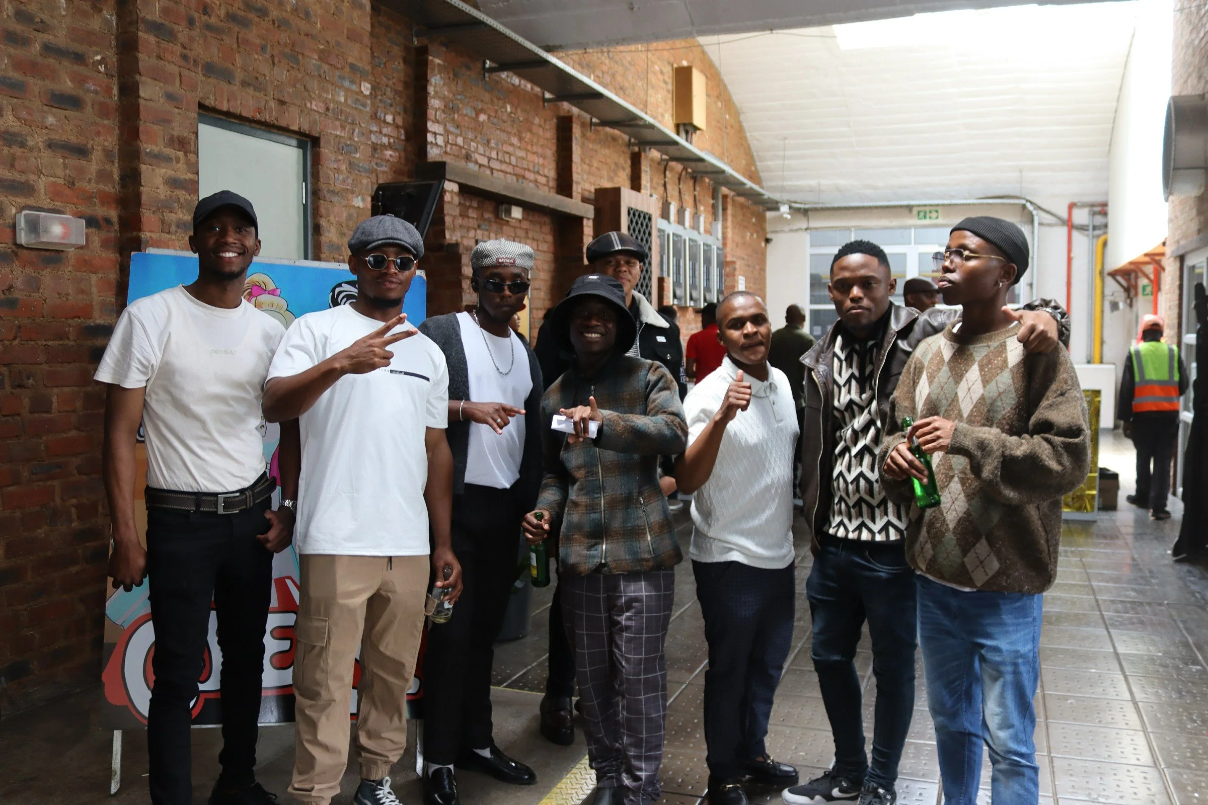 A group of seven young men standing inside a brick-walled building, posing for a photo. They are dressed casually, some with hats and sunglasses, and appear to be at a social event or gathering. There are people in the background and a colorful poste