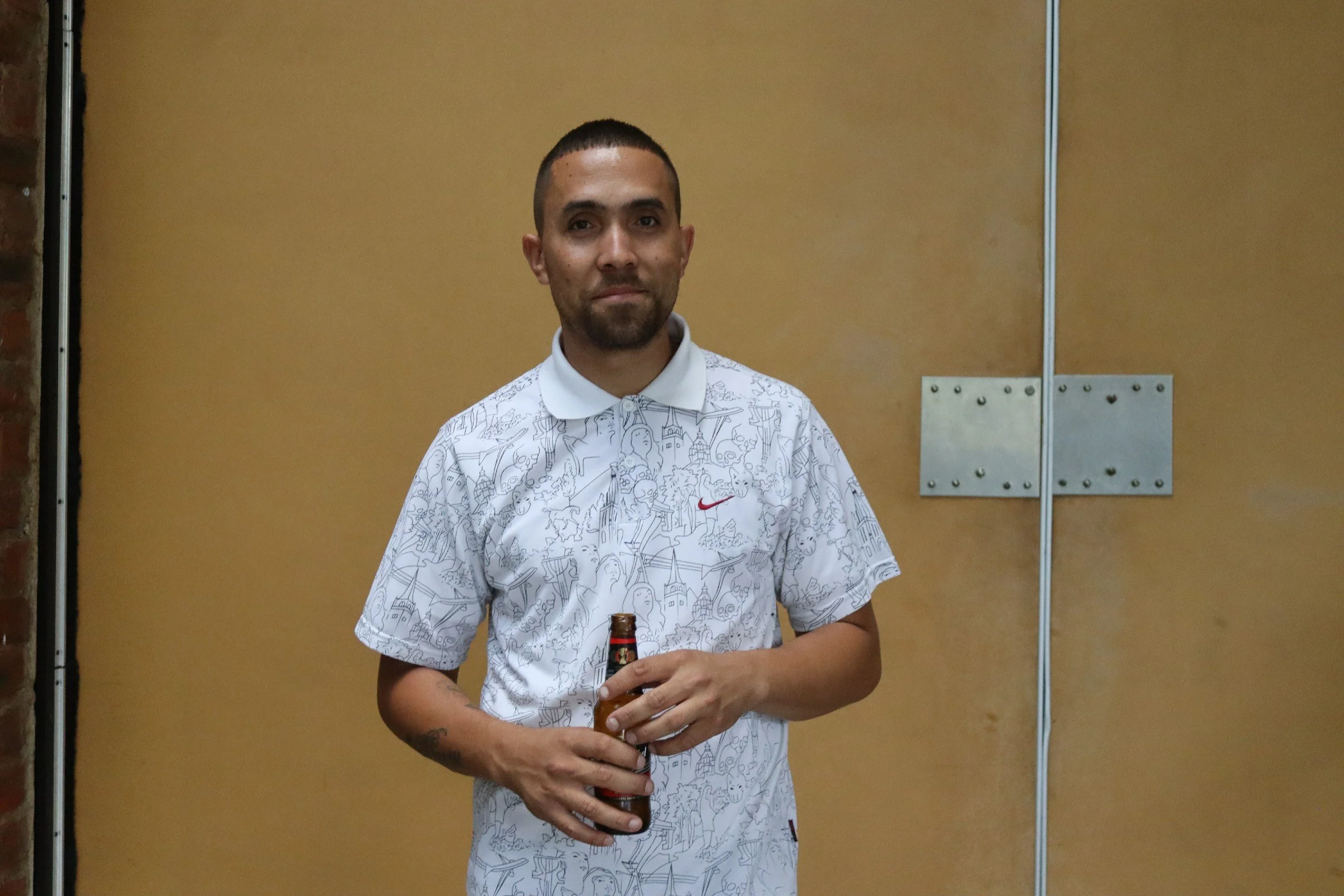 A man with short dark hair and a beard, wearing a white graphic shirt, holding a brown bottle, standing in front of a beige wall with metal door hinges.