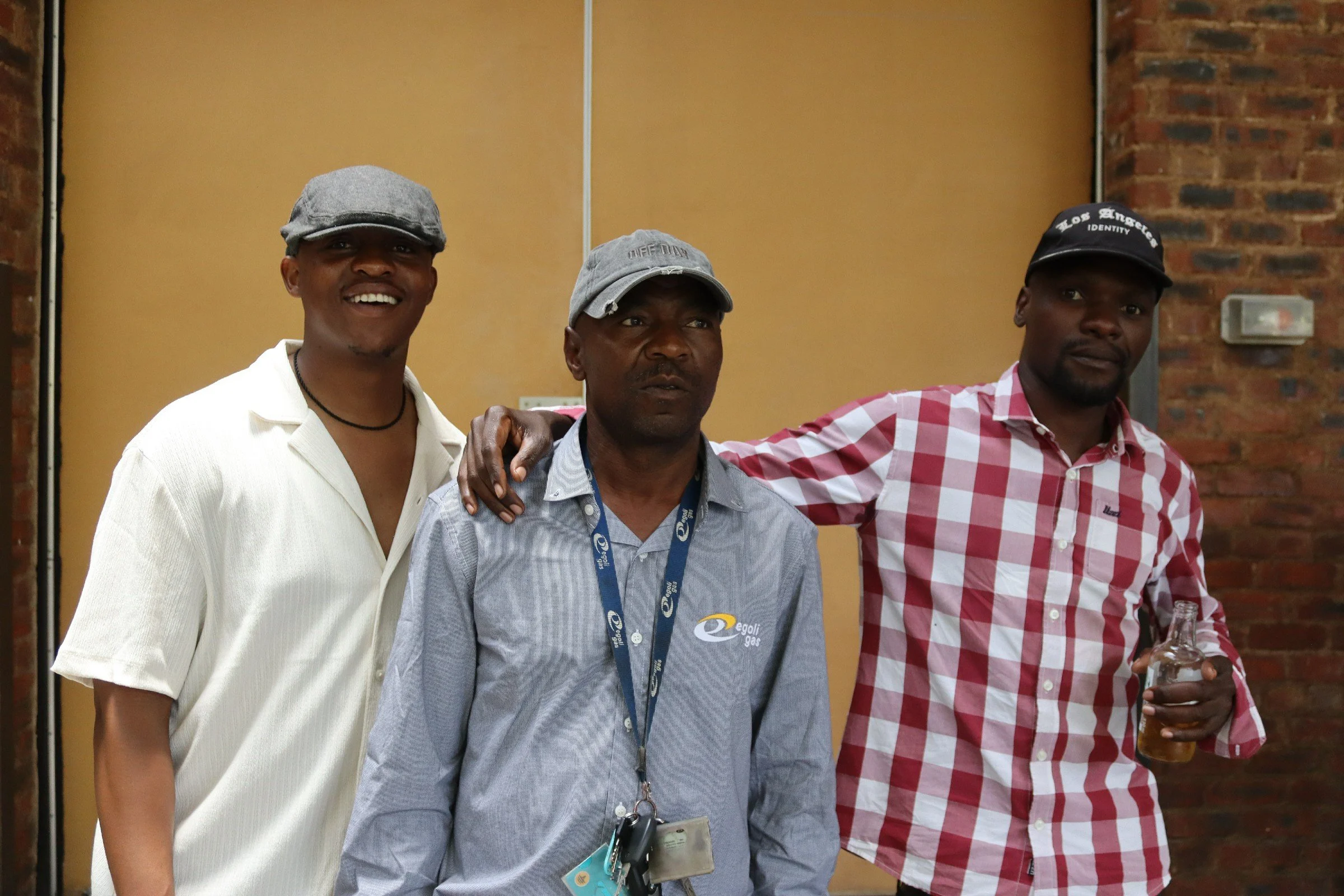 Three men smiling and posing for a photo indoors. The man on the left is wearing a gray cap and white shirt, the man in the middle is wearing a gray cap and a blue lanyard with a badge, and the man on the right is wearing a black cap and a red checke