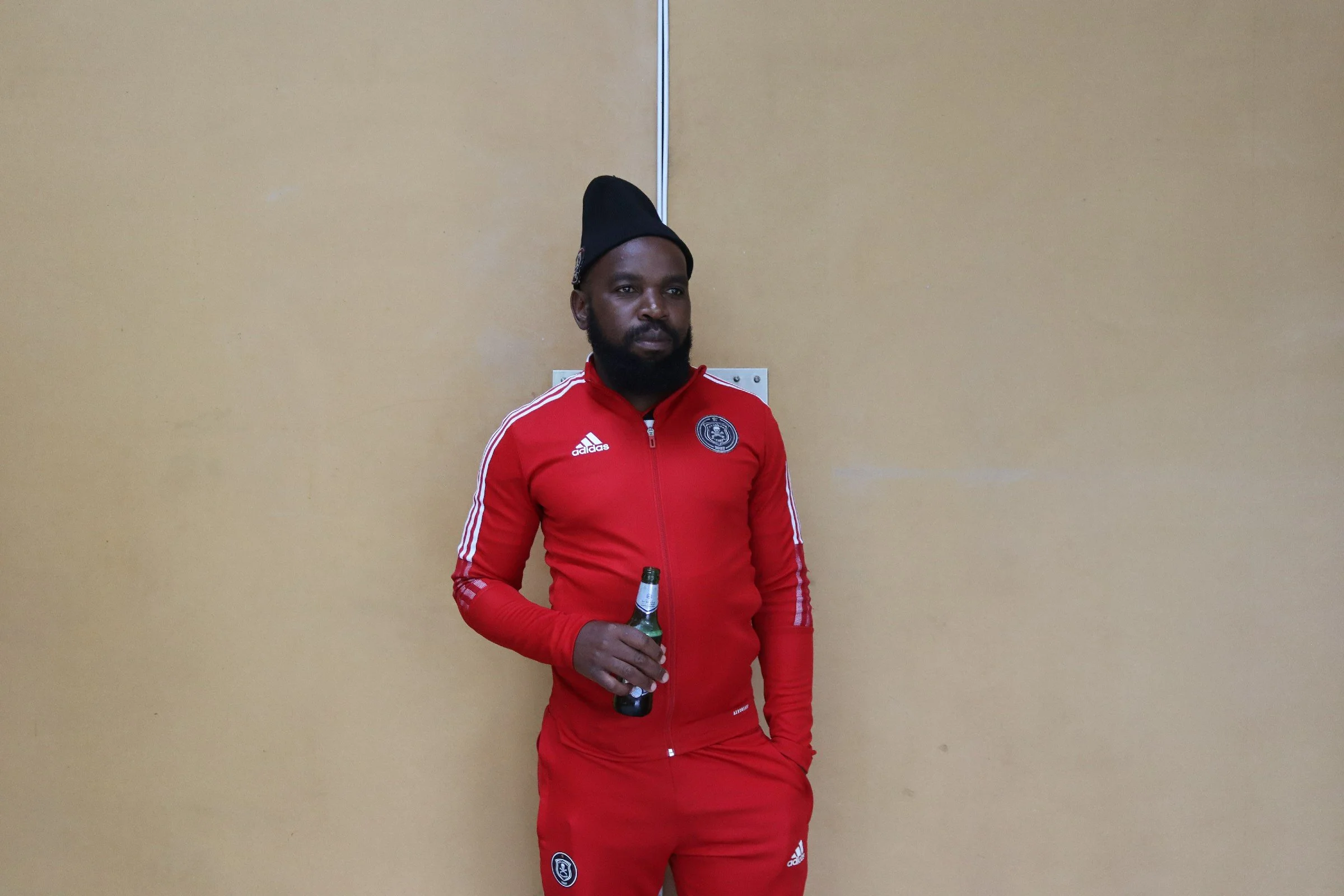 Man dressed in red sportswear holding a beer bottle, standing against a plain beige wall.