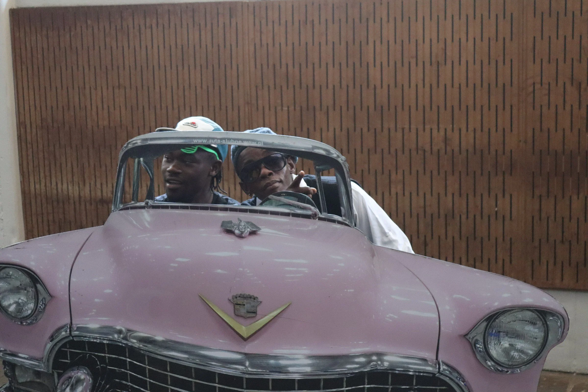 Two men sitting in a vintage pink Cadillac convertible, with a Cadillac emblem on the front and a gold V-shaped ornament, parked in front of a wooden wall.