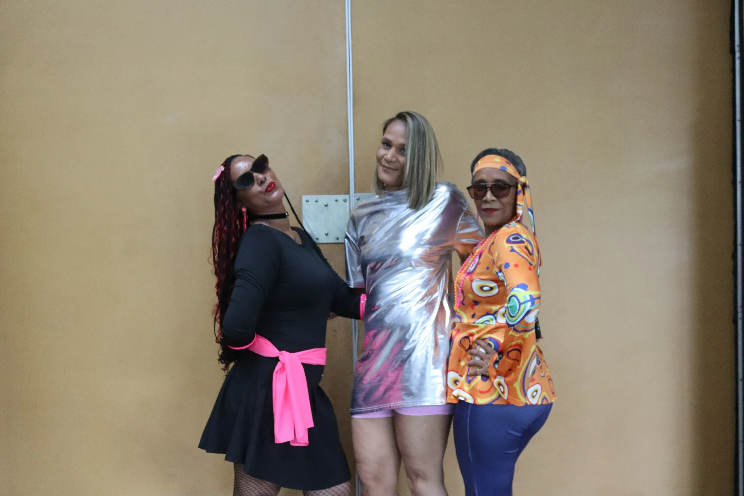 Three women dressed in colorful, fashionable outfits posing in front of a plain brown wall.