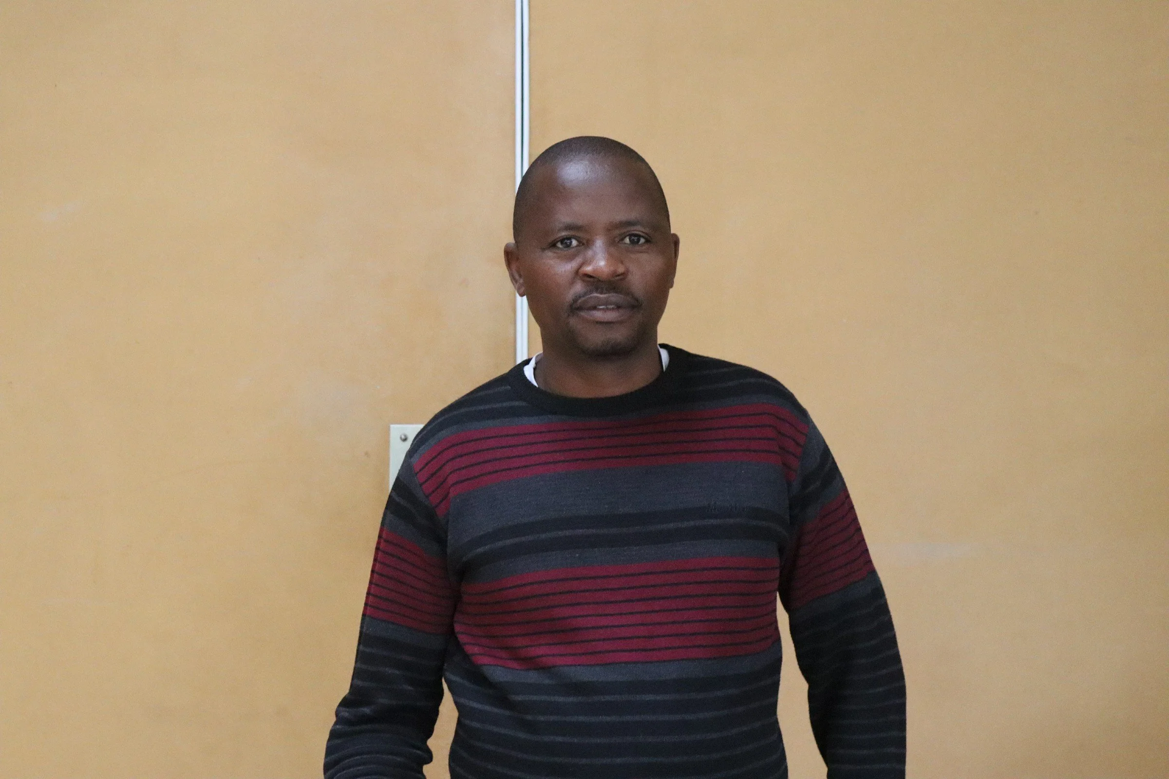 A man with a shaved head standing against a yellow wall, wearing a black, red, and gray striped sweater.