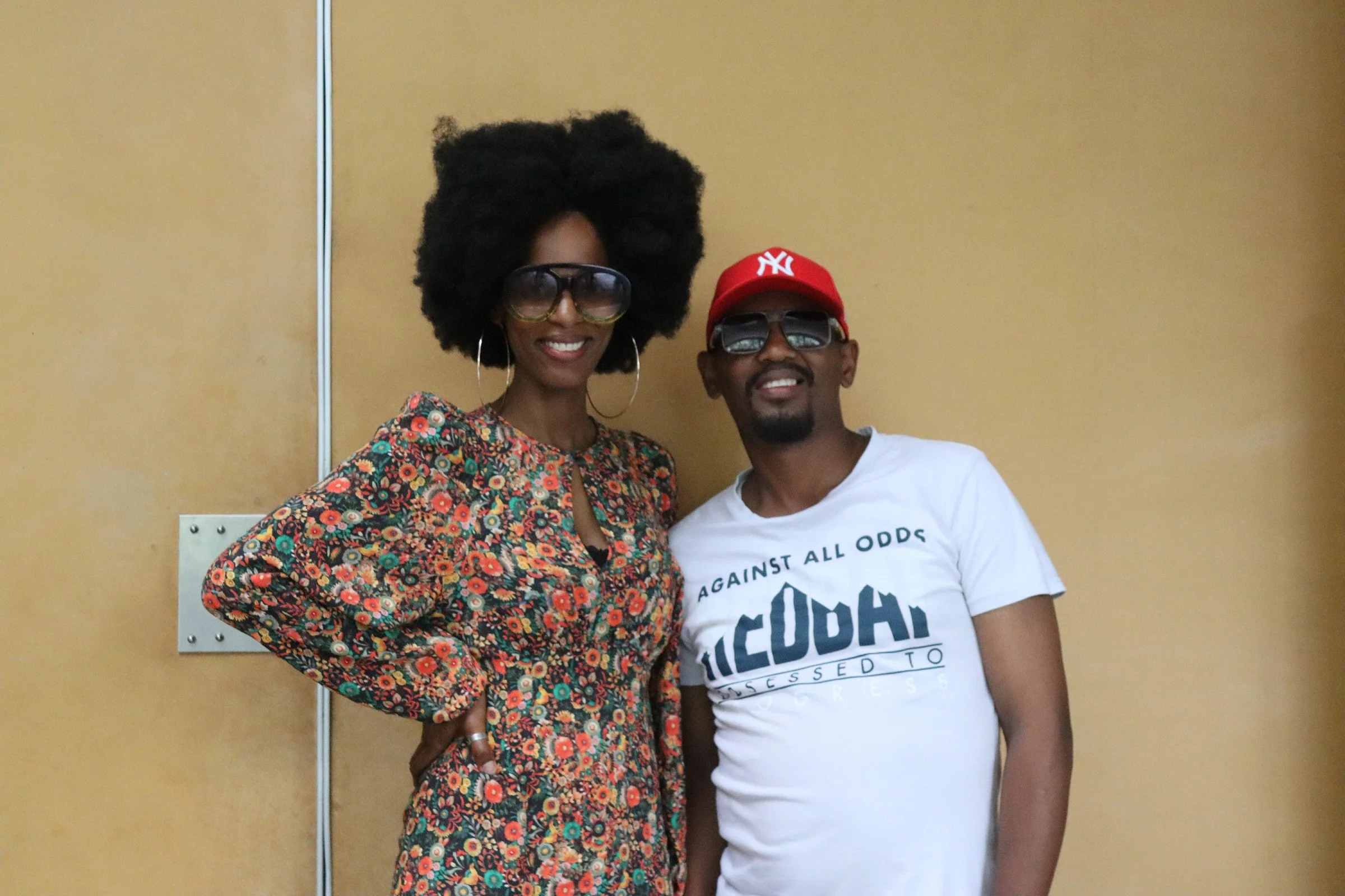 A woman and a man standing together in front of a yellow wall. The woman is wearing large glasses, hoop earrings, and a colorful floral dress. She has a big natural afro hairstyle. The man is wearing sunglasses, a red baseball cap, and a white shirt 