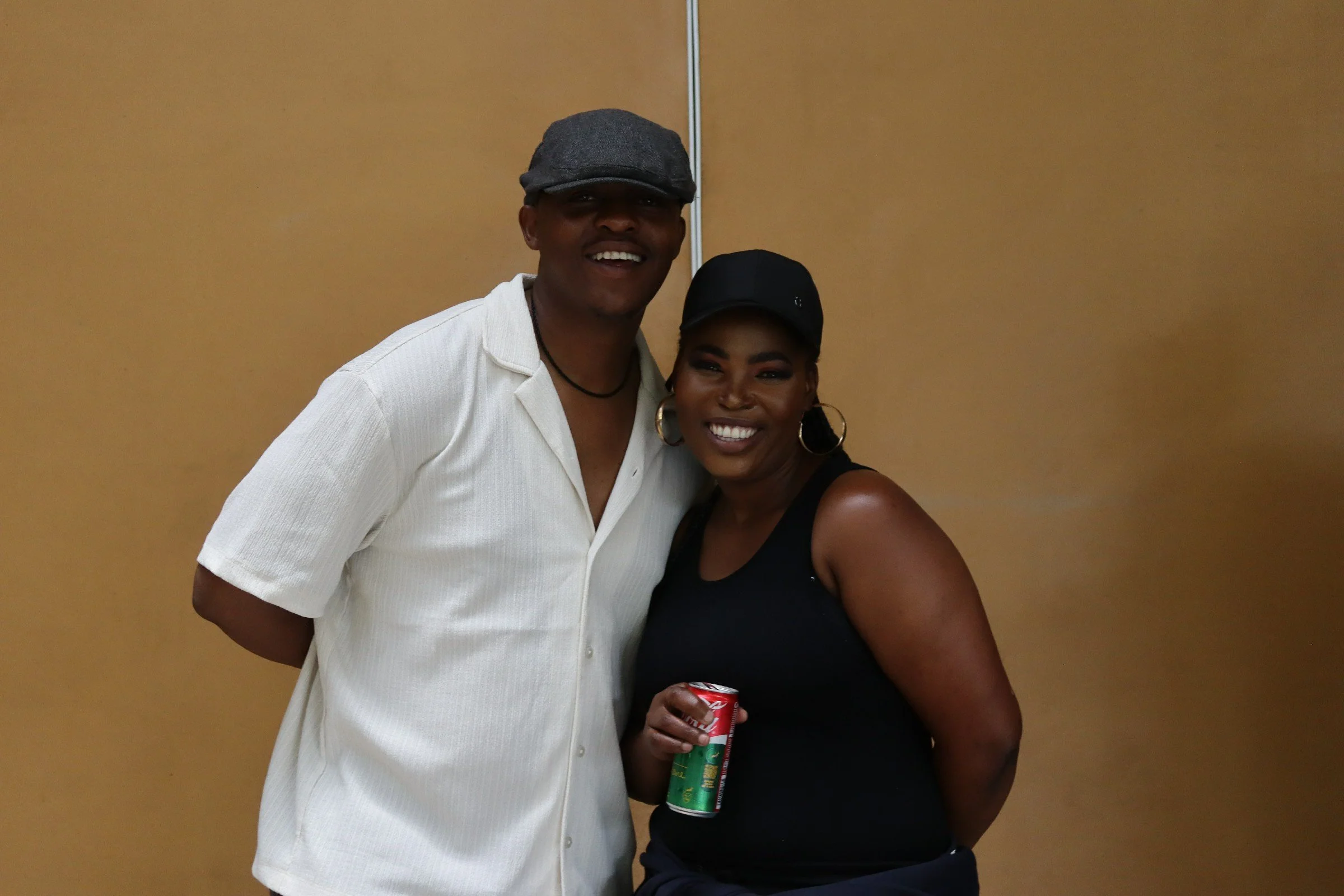 A man and woman smiling, standing close together against a plain tan wall. The man is wearing a gray cap and a white shirt, and the woman is wearing a black cap, black top, large hoop earrings, and holding a red soda can.