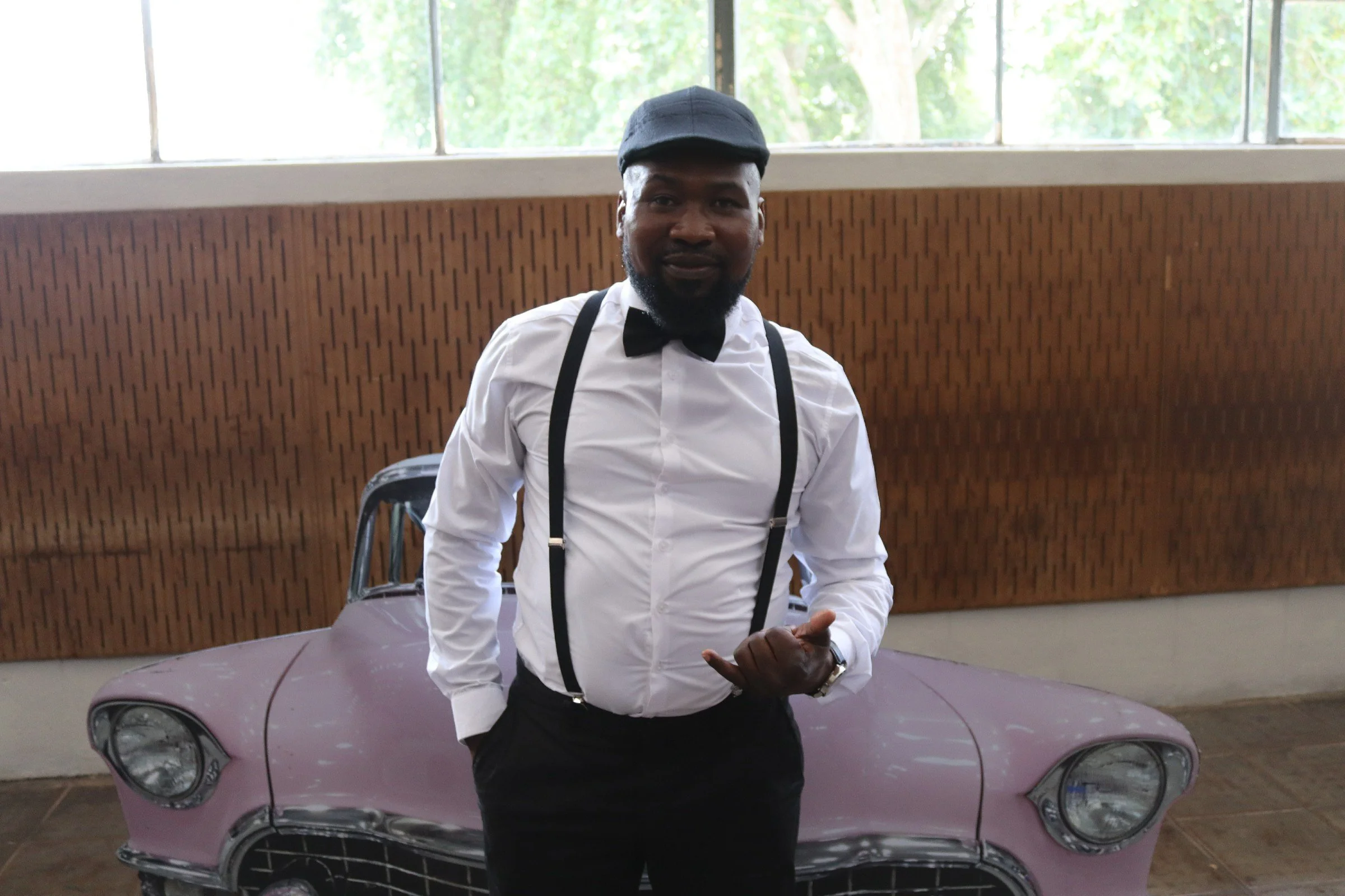A man dressed in a white shirt, black suspenders, and a black bow tie, wearing a black cap, stands in front of a vintage pink car inside a building with large windows and wood-paneled walls.