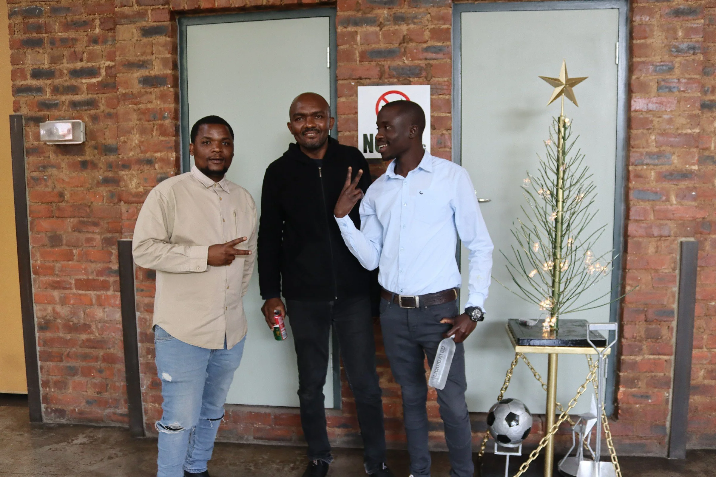 Three men posing in front of a brick wall with a Christmas tree decorated with a gold star on top. One man is holding a can of soda, and a small soccer ball is on the ground near the tree.