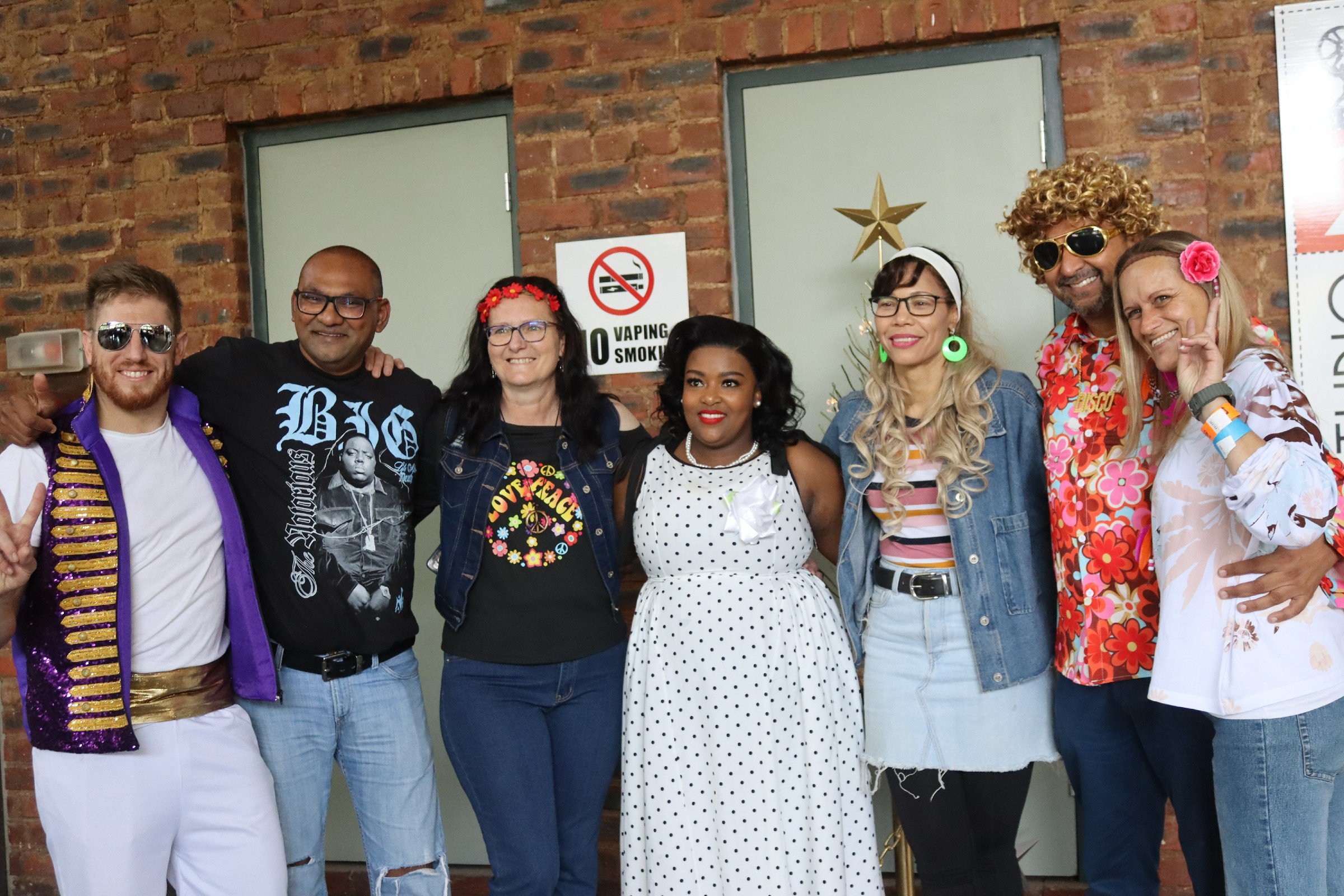 Group of seven diverse people dressed in colorful 70s-themed costumes posing together indoors against a brick wall.