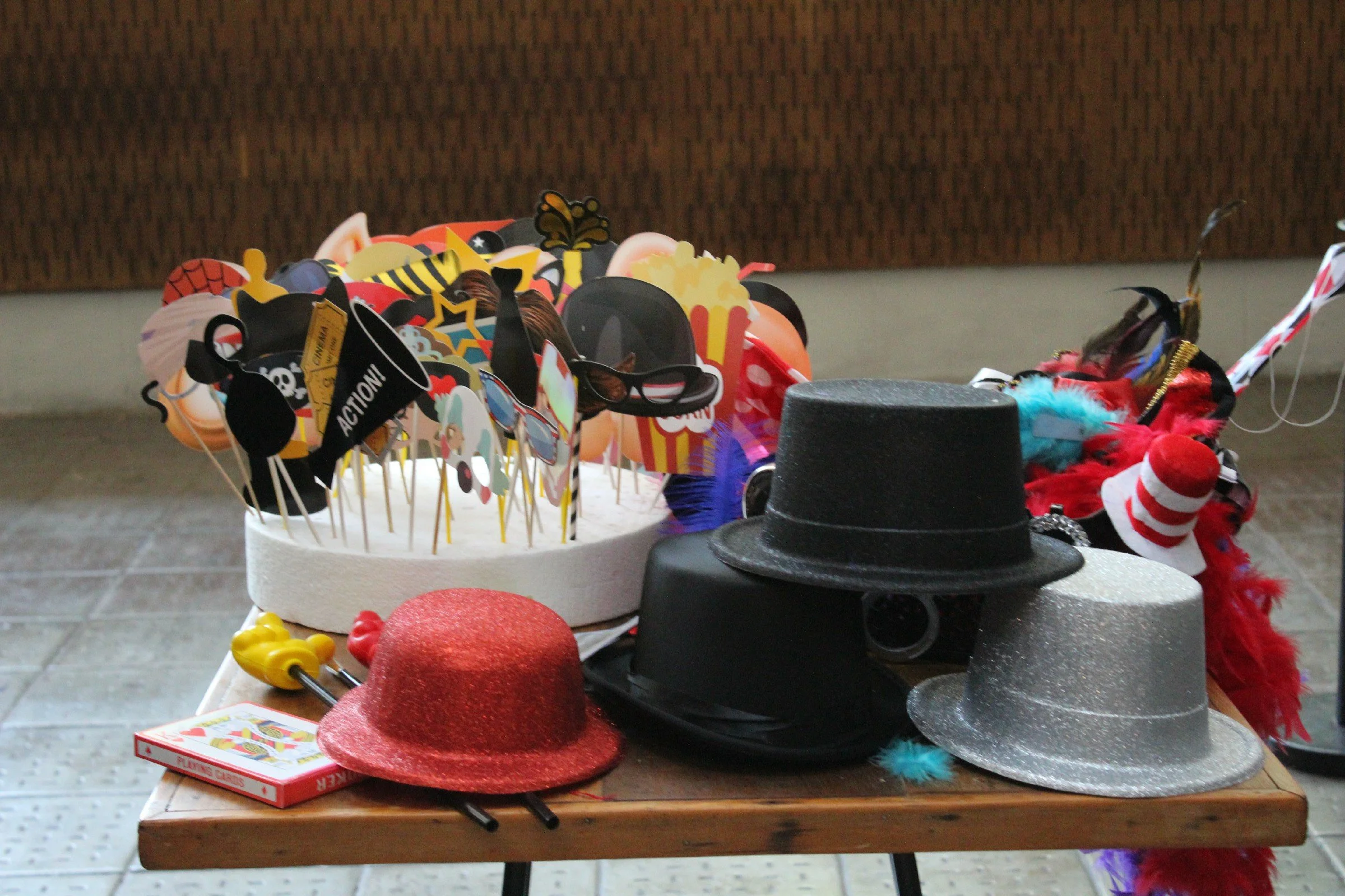 A table displaying various costume accessories such as hats, masks, and props, including a magician's hat, a red glittery hat, a silver glittery hat, and animal-themed items, with a box of playing cards nearby.