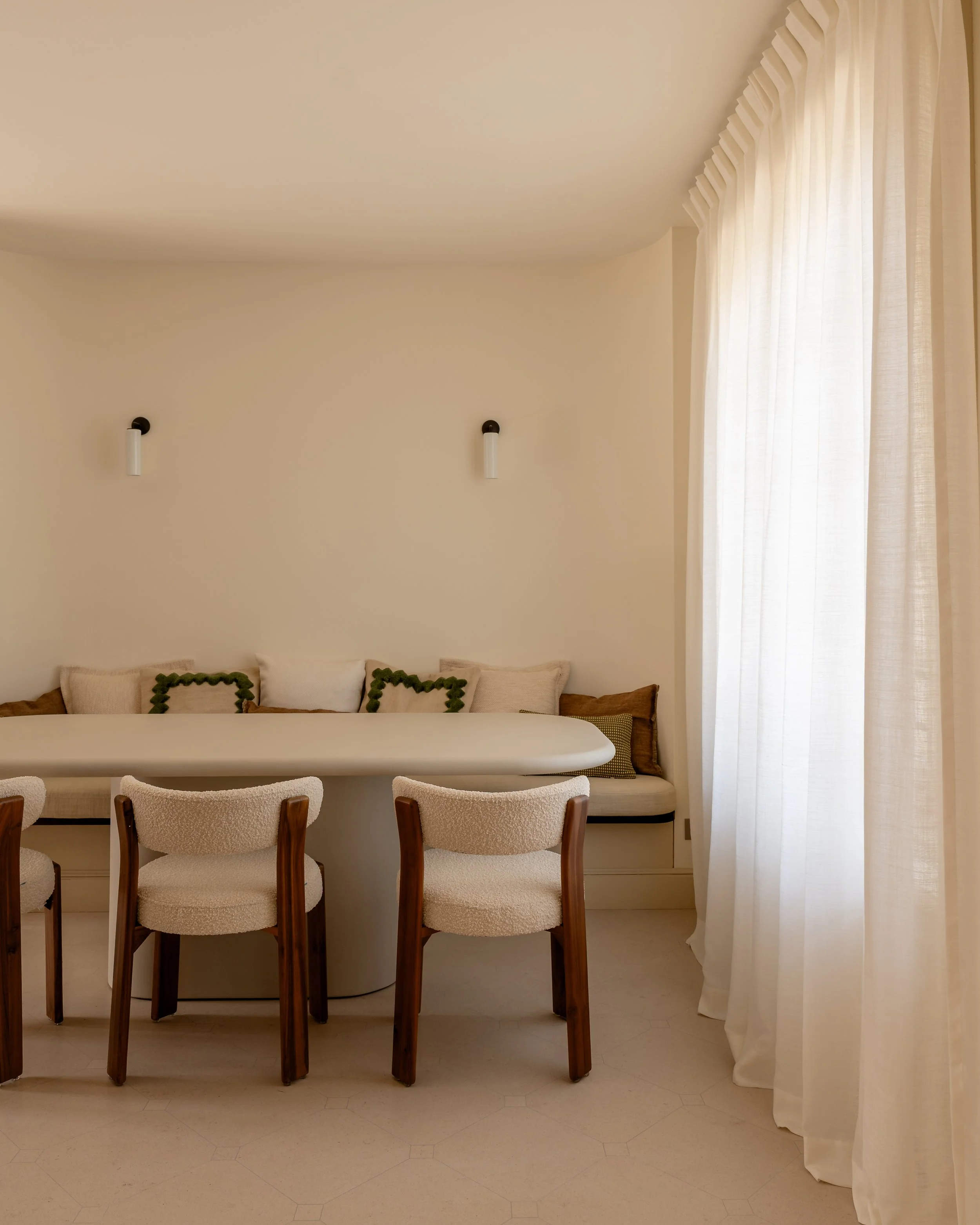 A dining area with a white round table, four chairs with wooden legs and cream-colored upholstered seats, a beige sectional sofa with decorative pillows, wall-mounted lights, and large windows with sheer white curtains allowing natural light.