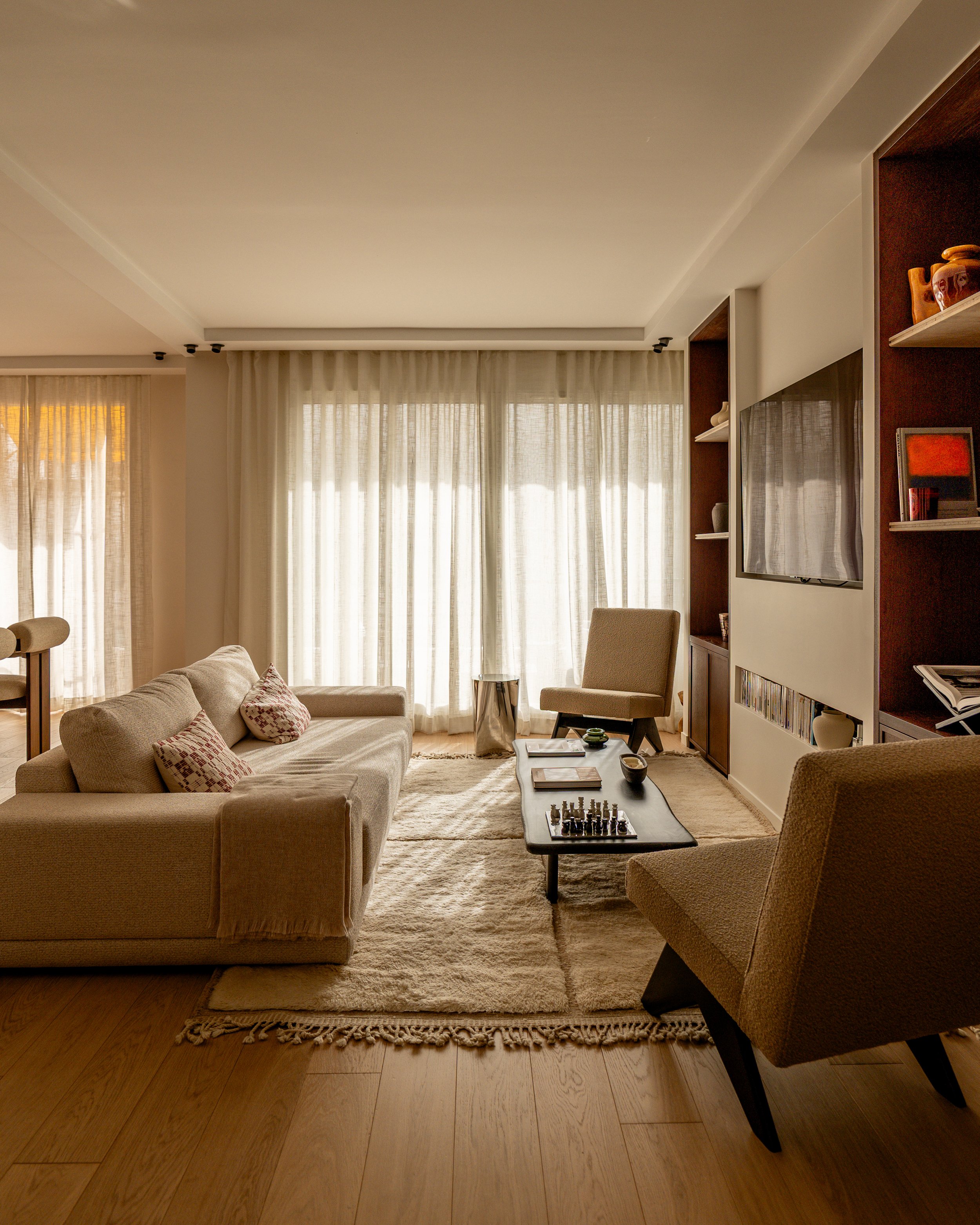 Living room with beige sofas, a coffee table with a chess set and books, a cream armchair, a beige rug, large windows with sheer curtains, and built-in shelves with decorative items and books.