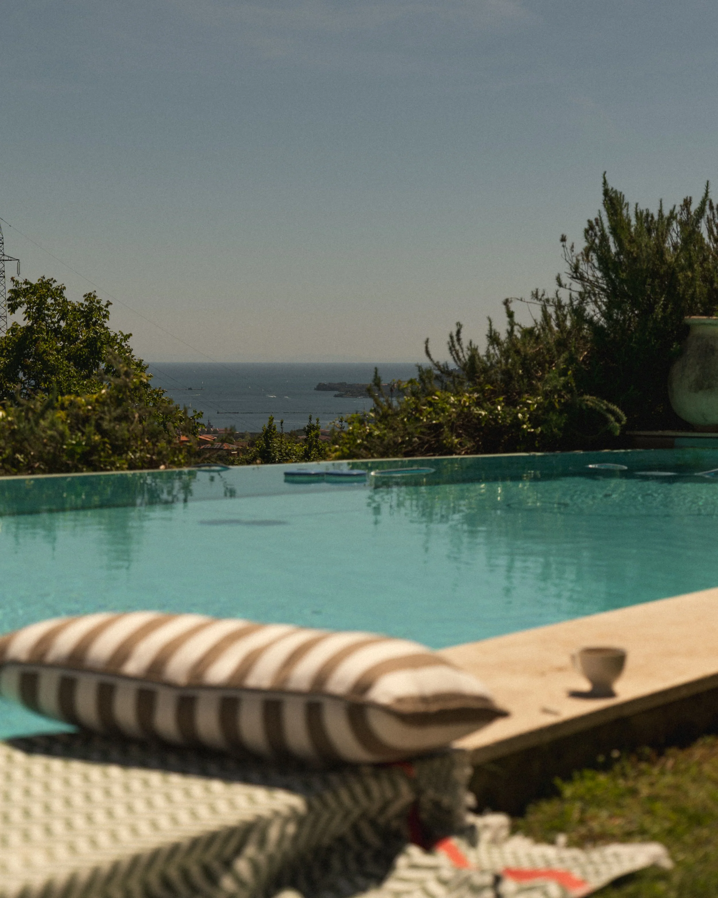 A view of a swimming pool with a striped lounge chair in the foreground and lush green trees and a body of water in the background, possibly an ocean or a lake.