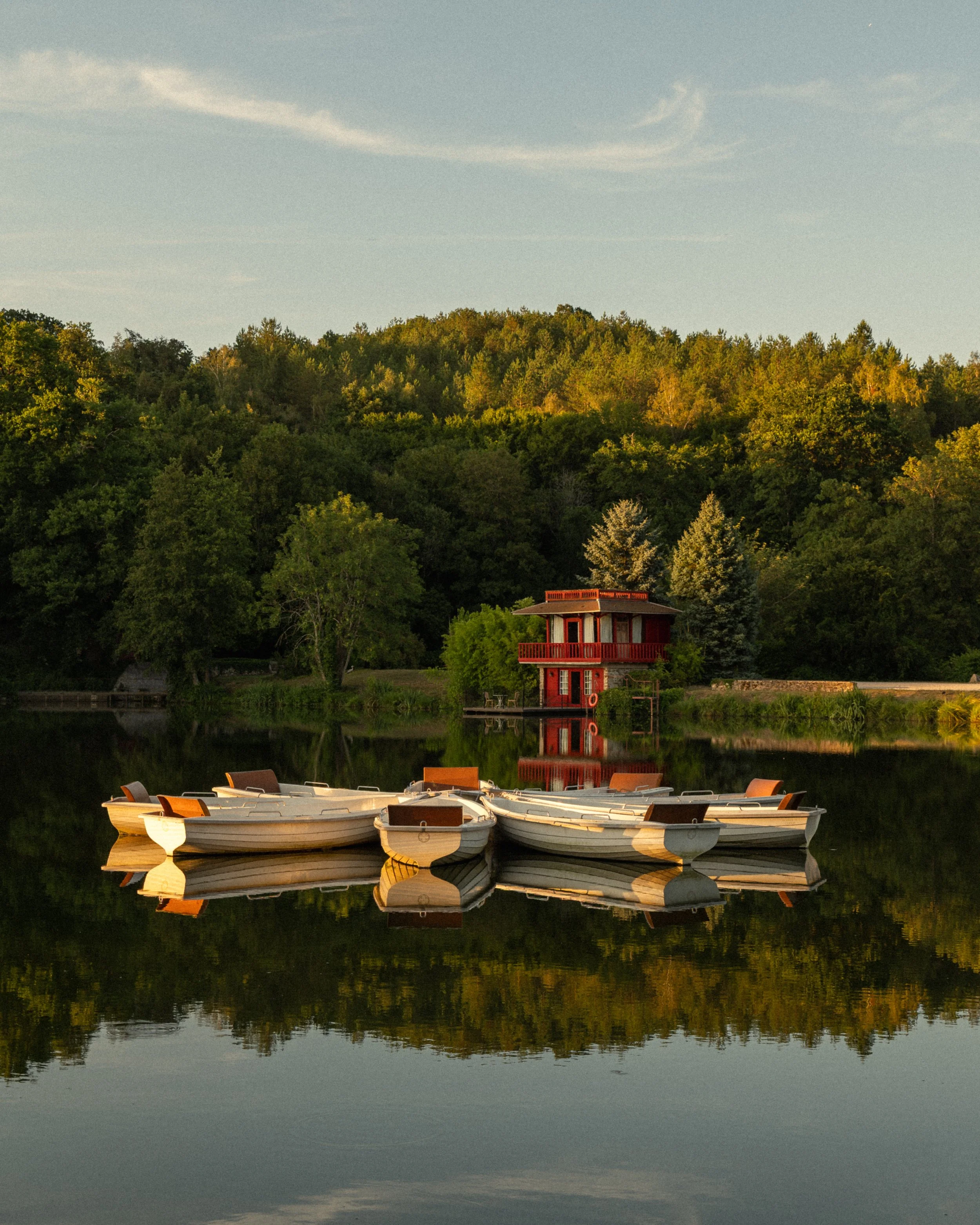 Rowboats docked by a calm lake, with a red house on stilts and a lush green hill in the background, all reflected in the water.