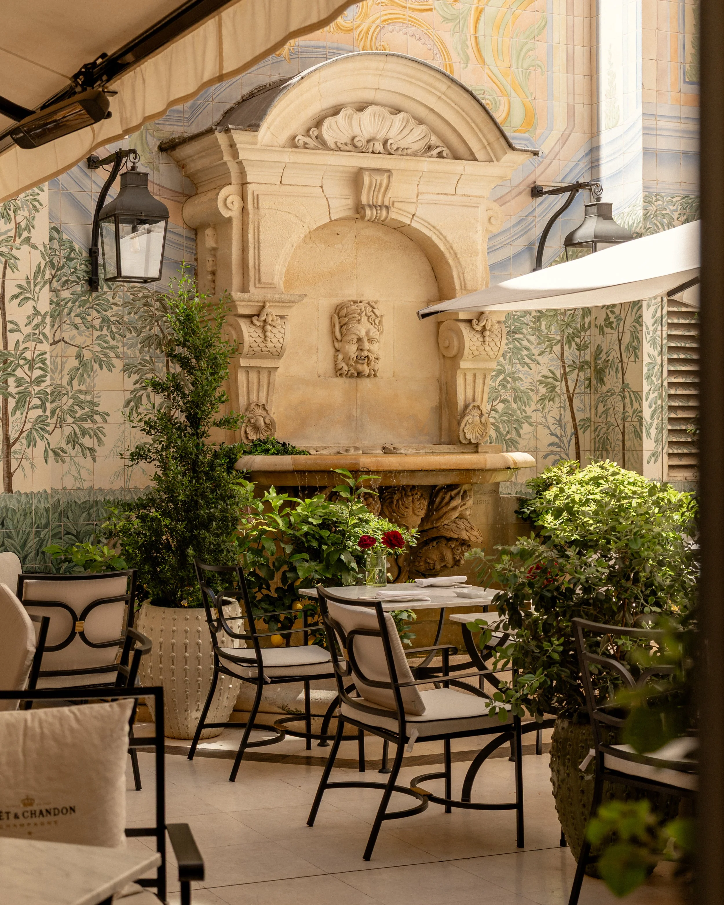 Elegant outdoor courtyard with a stone fountain featuring a carved lion's face, surrounded by lush green plants and chairs; decorative tiled wall with floral and geometric patterns; black wrought-iron lamps and a beige canopy overhead.