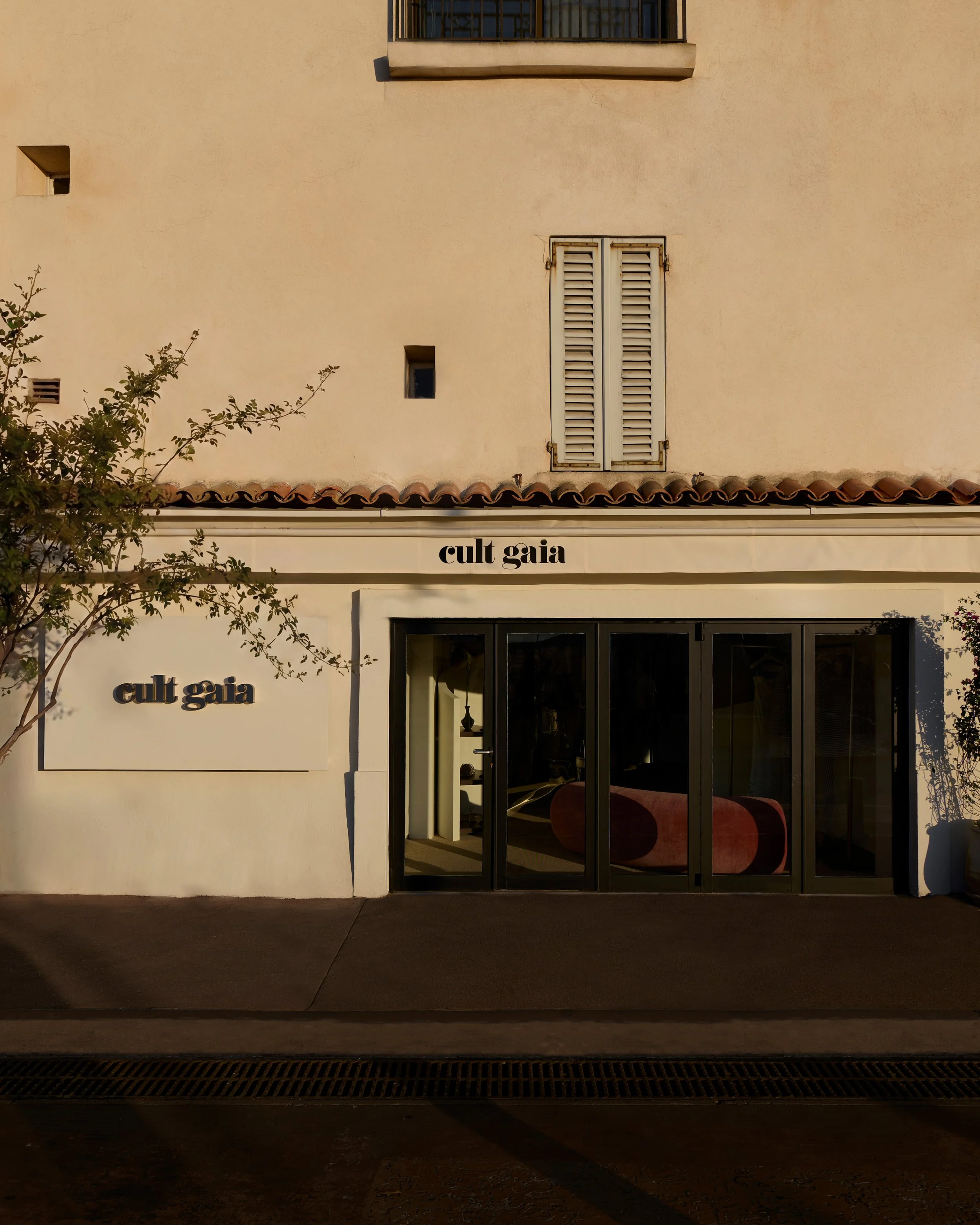 A beige building with small windows and a shuttered window on the second floor. The storefront has a sign that reads 'cult Gaia' with large glass doors below. There are some tree branches on the left side of the image, and the ground is dark and text