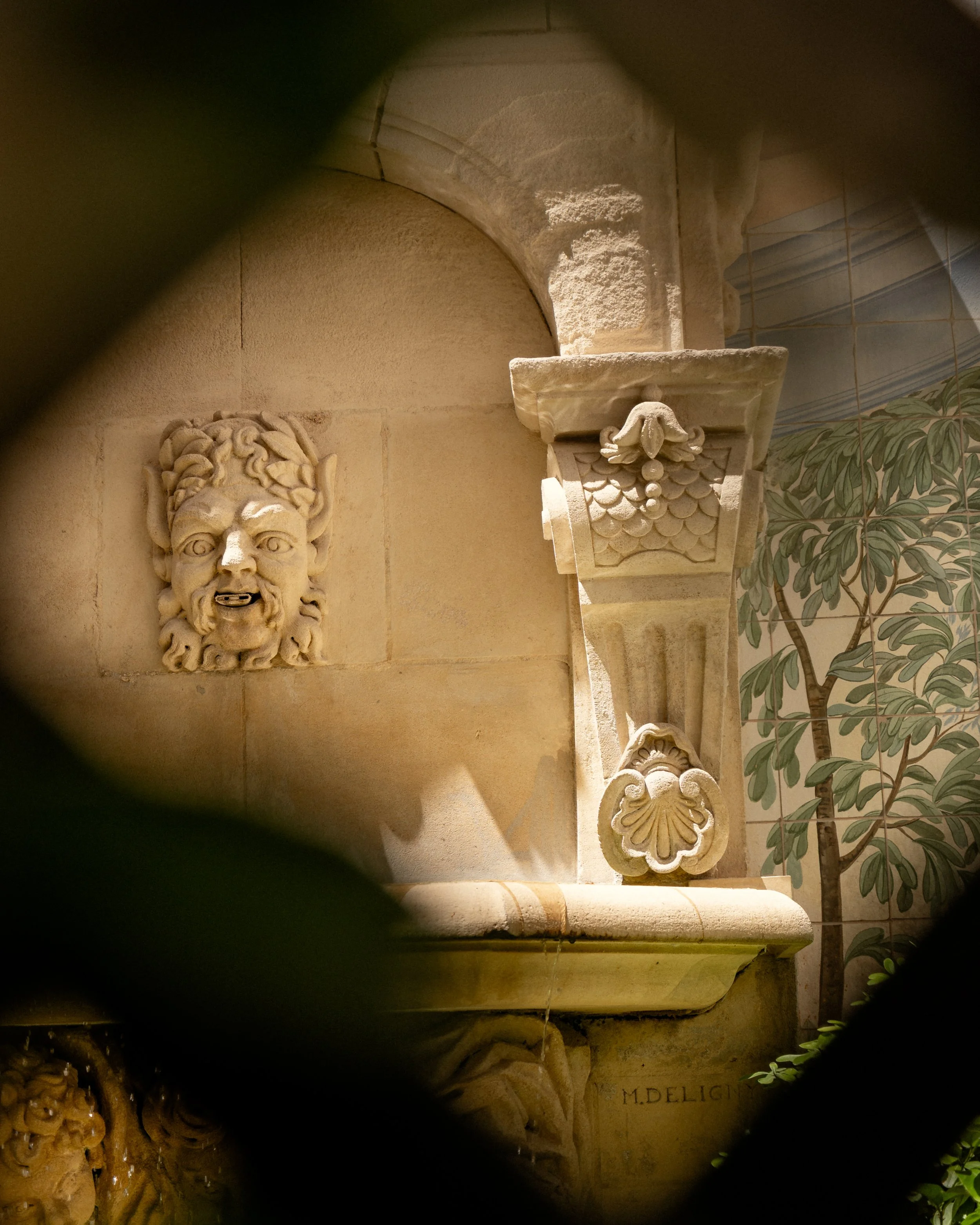 Close-up view of a decorative stone carving and architectural details on a beige stone wall, including carved faces and intricate patterns, with a potted plant with green leaves partially visible on the right.