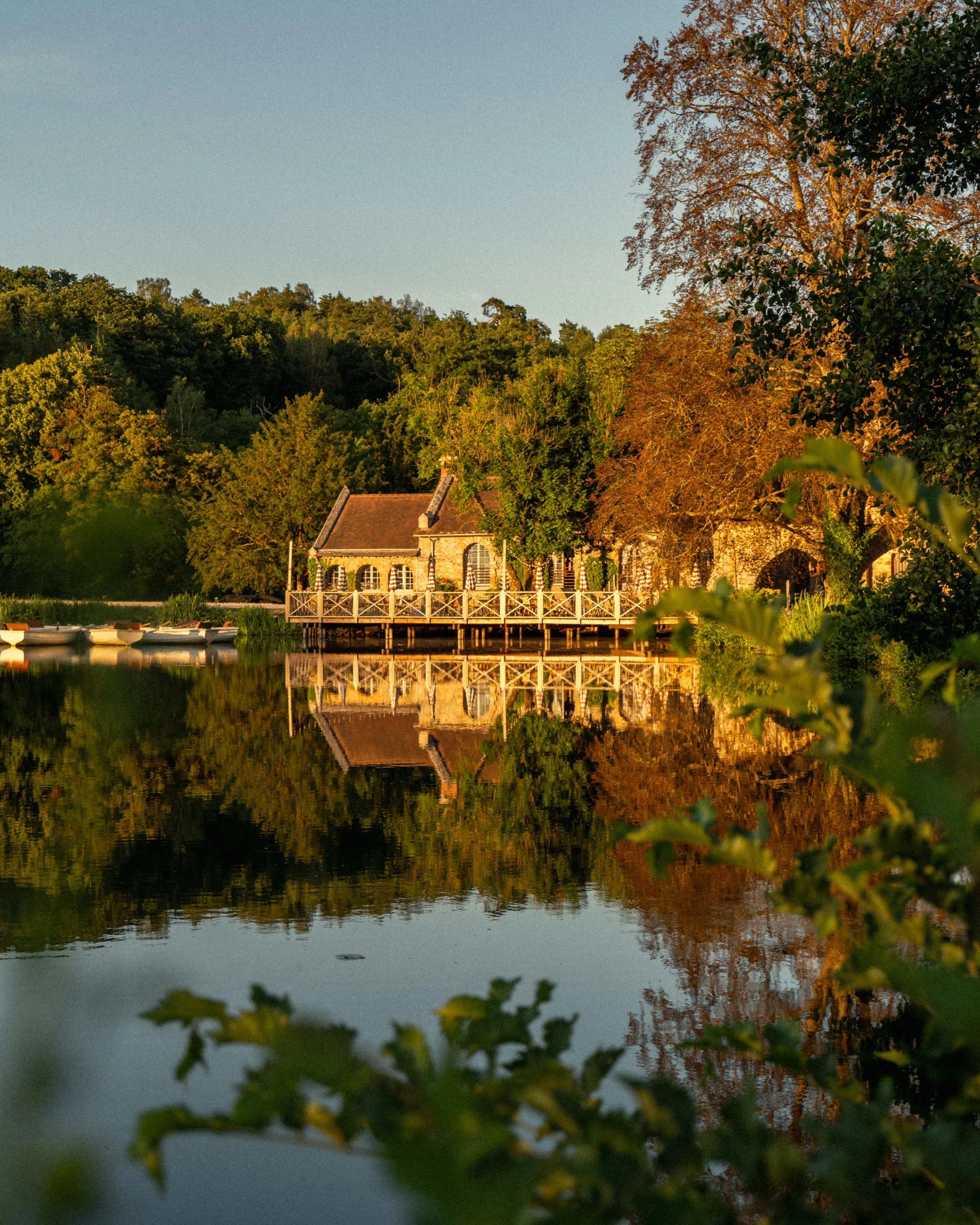 A peaceful lakeside scene with a house on the shore, surrounded by lush trees displaying fall foliage, and their reflections visible on the calm water. The sky is clear and the lighting suggests either early morning or late afternoon.