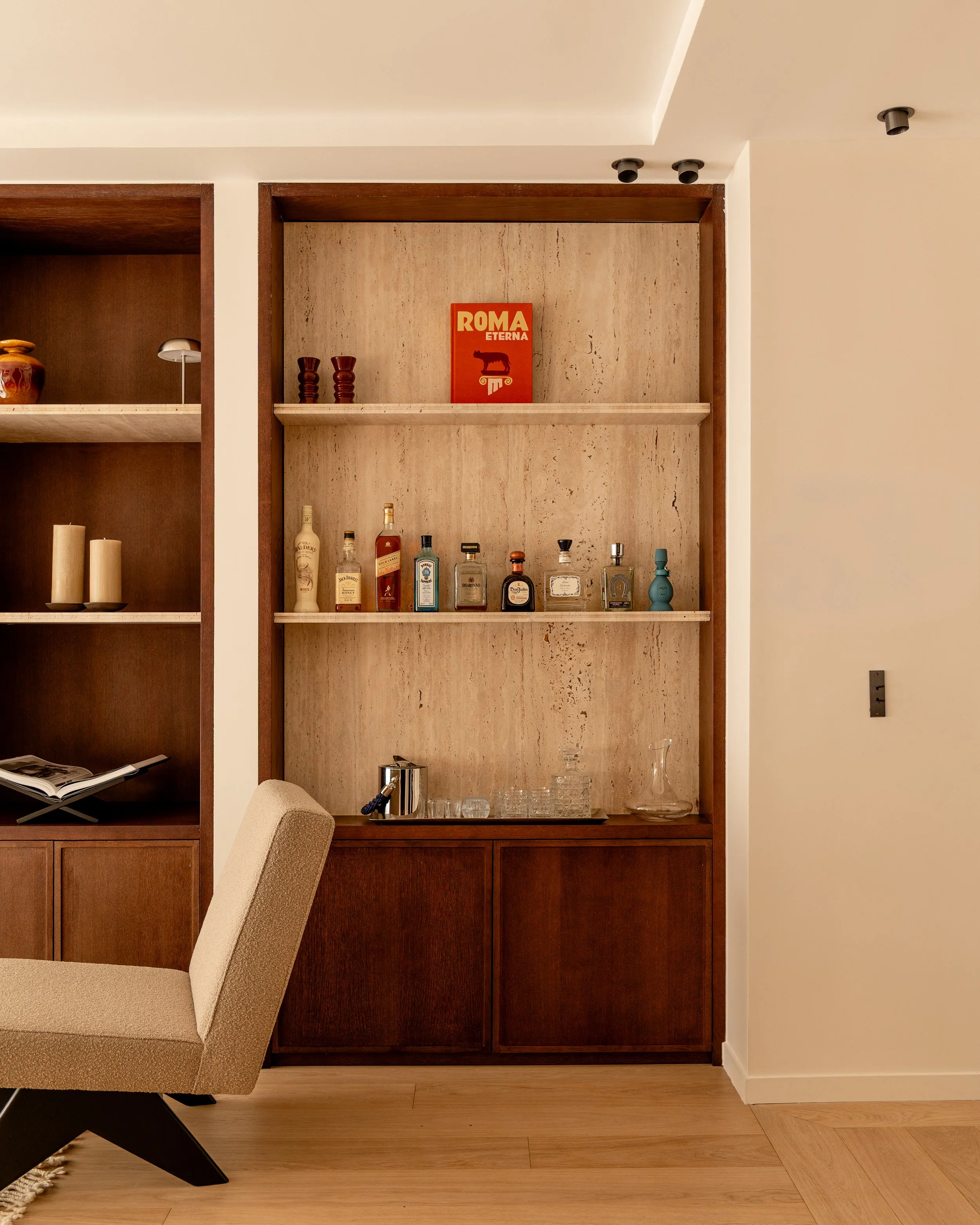 A built-in wooden bar shelf with various alcohol bottles and glasses, a small sculpture, and a framed art piece with the text "ROMA ETERNA" and a stylized wolf head, set against a beige wall with decorative trim and multiple ceiling lights.