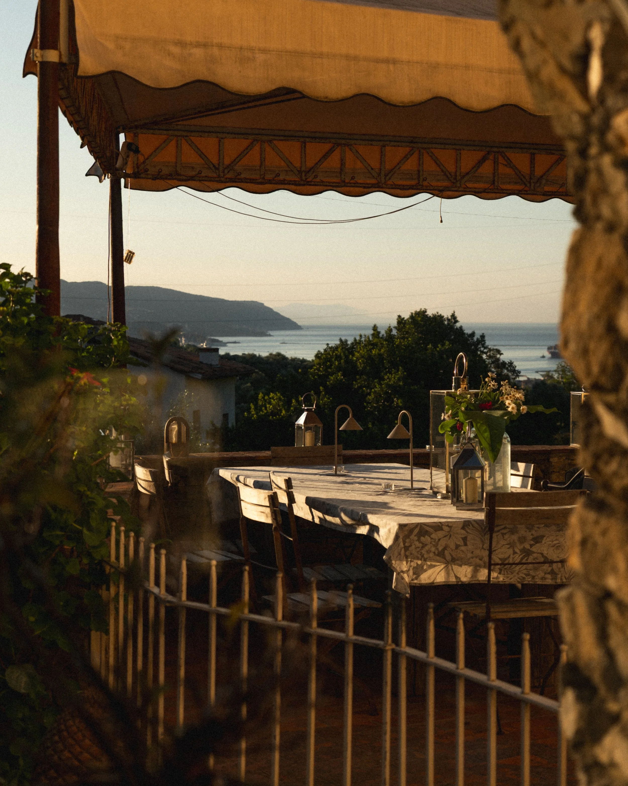 A view from a balcony or patio overlooking a scenic body of water with hills in the distance during sunset or sunrise. There is a table with a tablecloth, lanterns, and plants in the foreground. A yellowish awning provides shade, and a wooden fence s