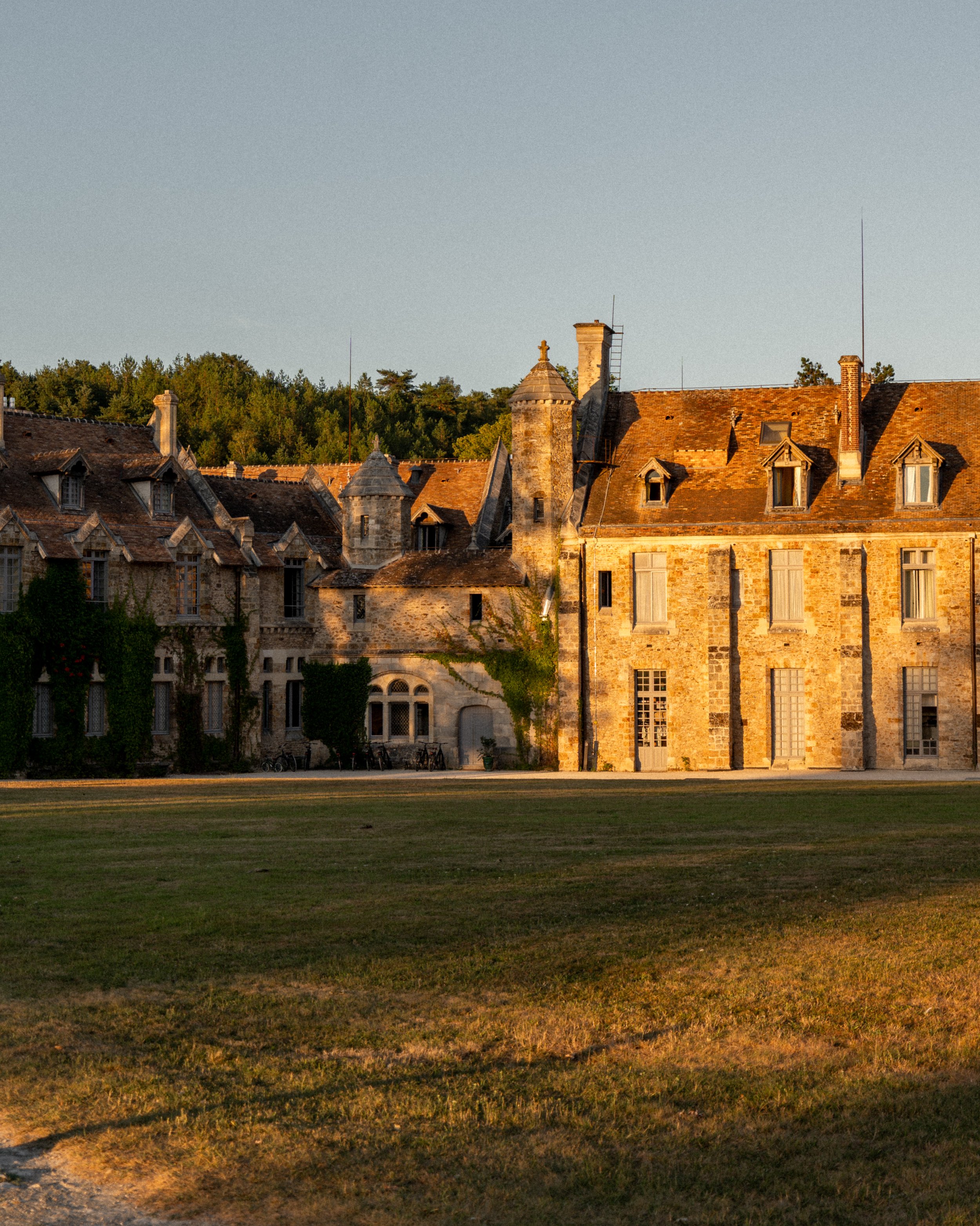 A historic stone building with multiple chimneys, dormer windows, and a tiled roof, surrounded by a grassy lawn and set against a backdrop of trees and a clear sky at sunset.