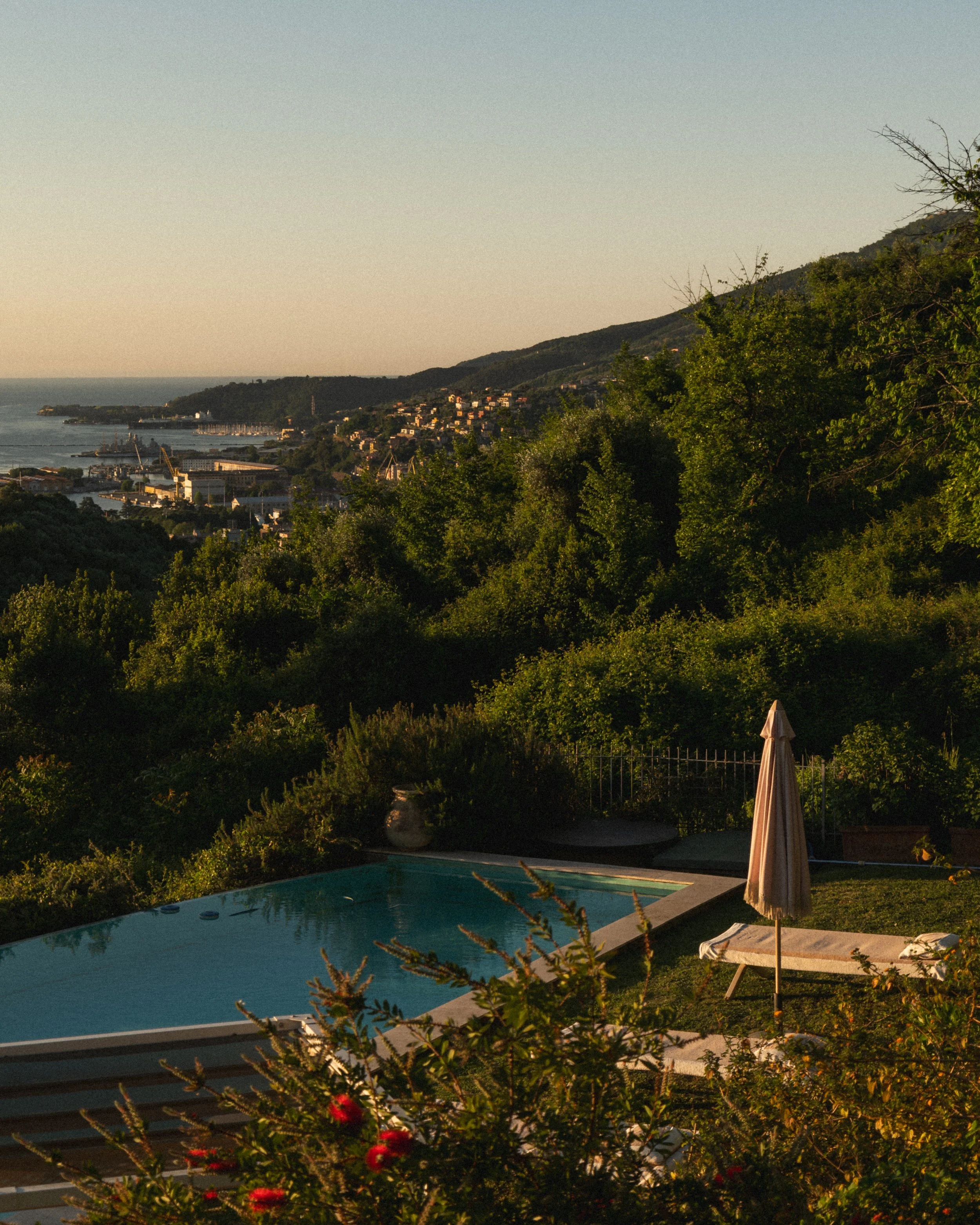 A backyard with a swimming pool, a patio umbrella, a lounge chair, lush green trees, and a scenic view of a hilly coastline with houses and a body of water, captured during sunset.