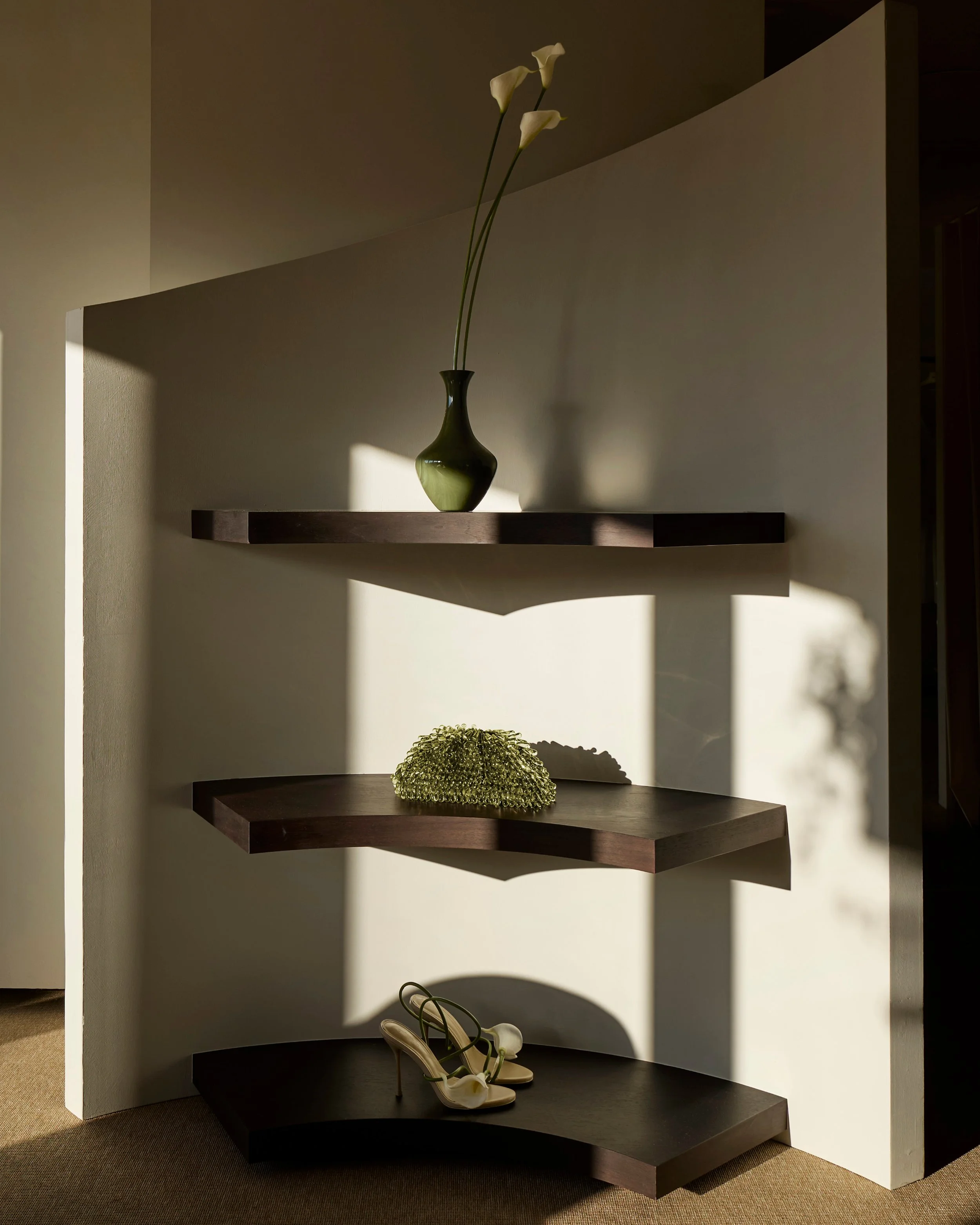 A dark green vase with white calla lilies on a curved dark wooden shelf against a white wall, topmost shelf empty, bottom shelf with a pair of white high-heeled shoes with flower details.