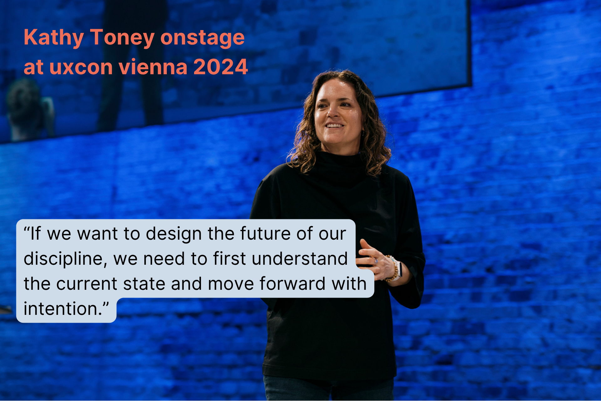 Kathy Toney on stage at Uxcon Vienna 2024, with a blue brick wall background, speaking into a microphone, wearing a black top and jewelry.