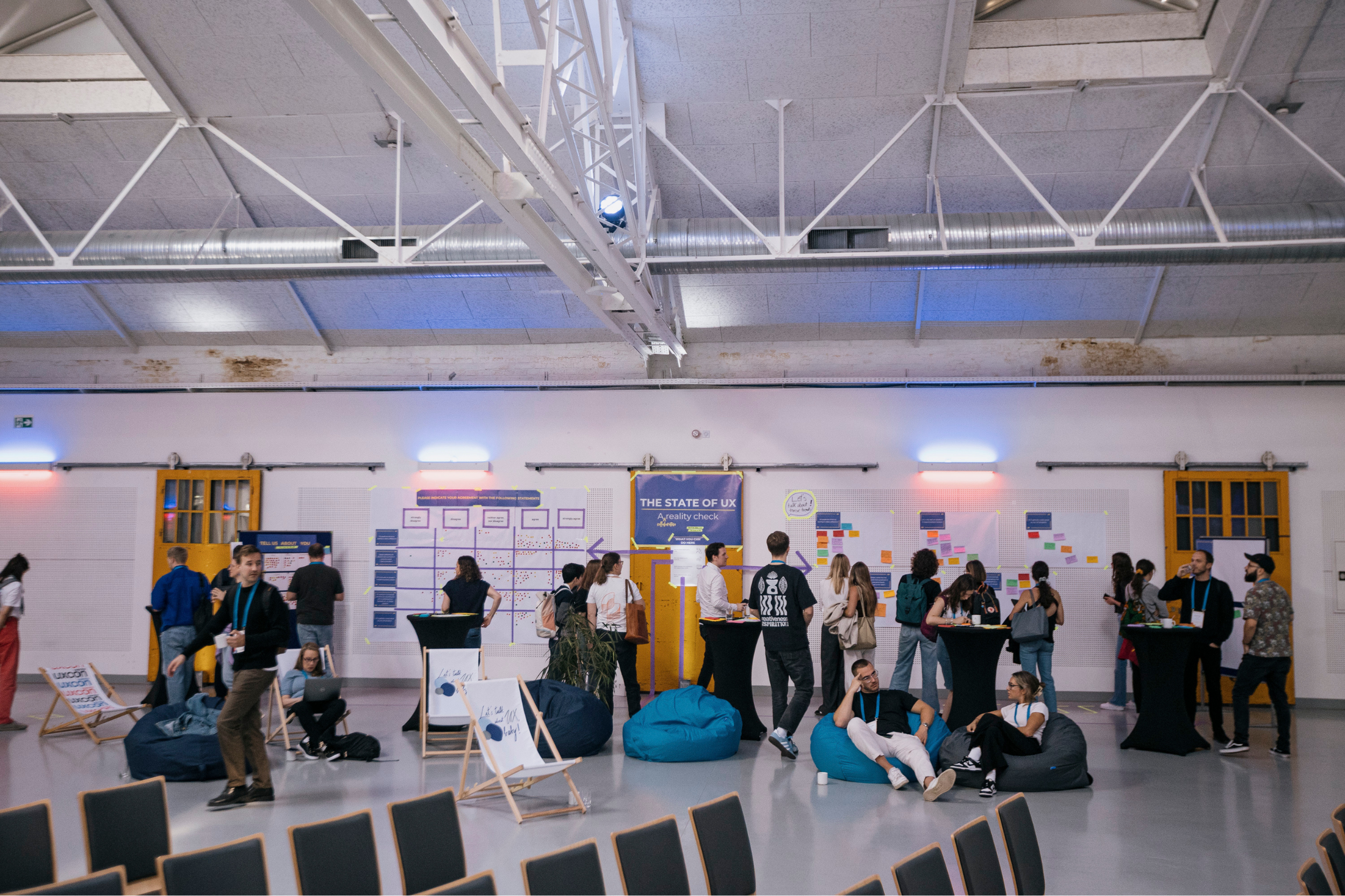 People gather at an indoor conference or event space with informational displays and seating. The ceiling shows metal beams, ductwork, and lights, while the walls have posters and screens.