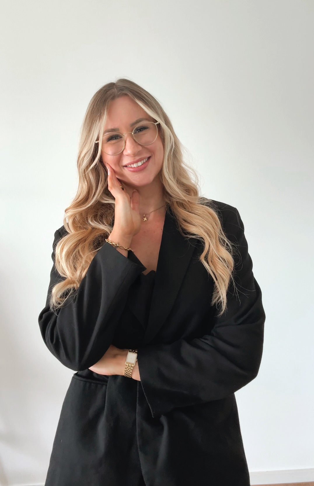 Eine Frau mit langen, gewellten blonden Haaren, Brille und einem schwarzen Blazer, die lächelnd in die Kamera schaut, vor einem weißen Hintergrund.