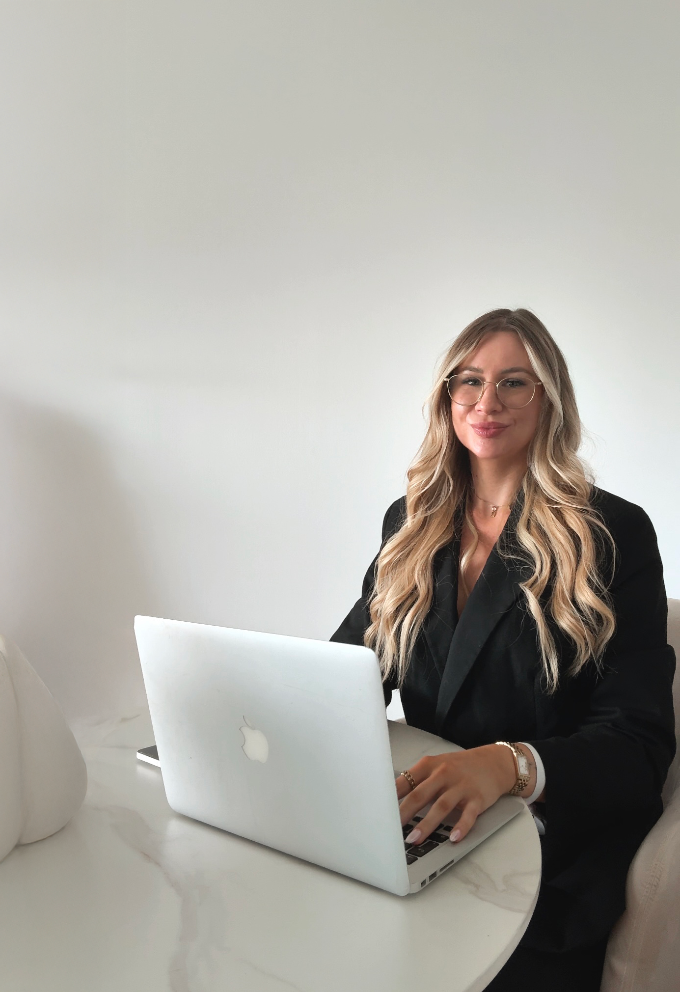 Eine Frau mit langen Blondhaaren, Brille und dunkler Kleidung sitzt an einem weißen Tisch vor einem offenen MacBook, während sie an einem Computer arbeitet.