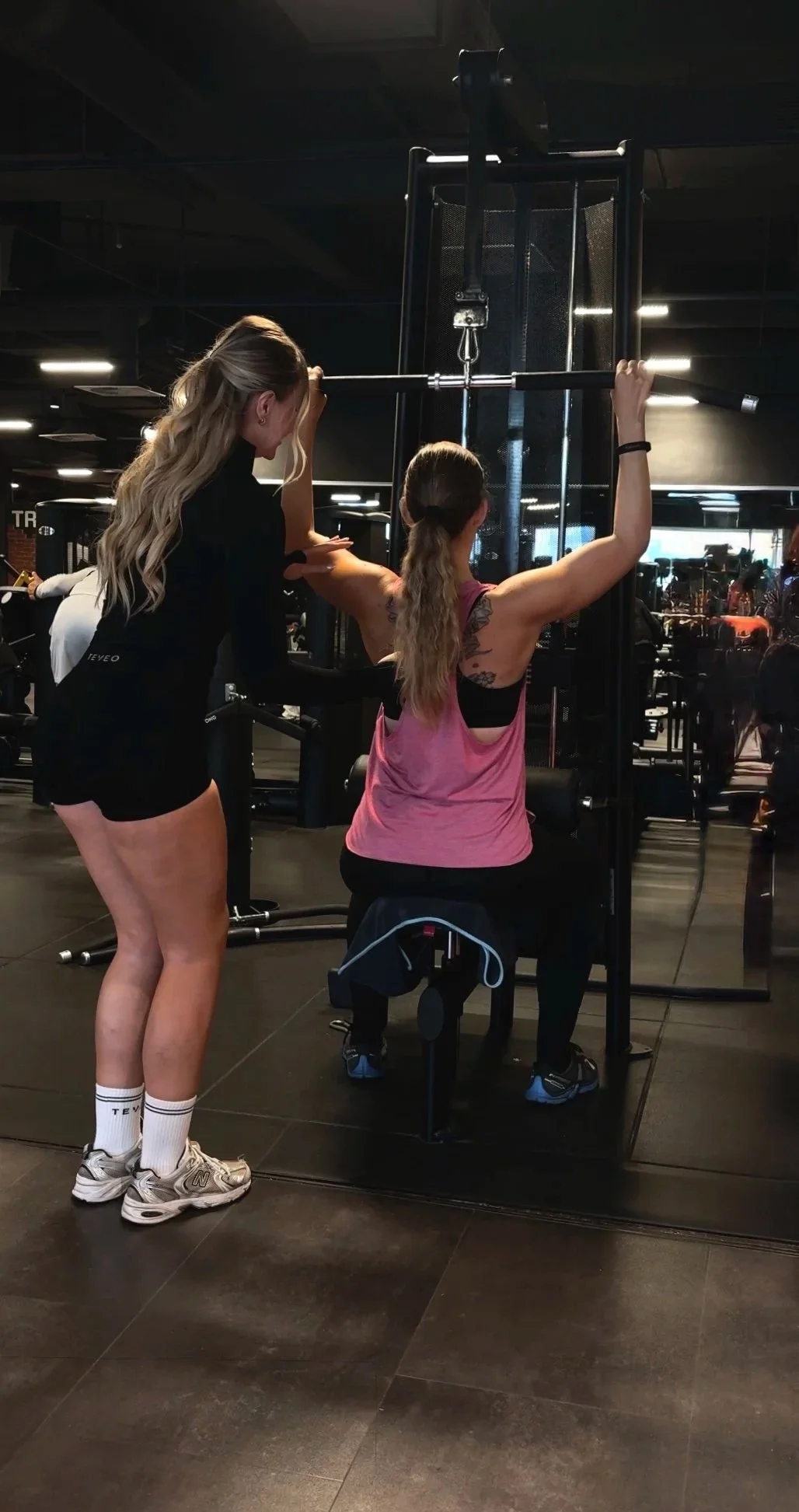 Zwei Frauen im Fitnessstudio, eine ist beim Krafttraining mit einer Maschine, die andere assistiert und beobachtet.
