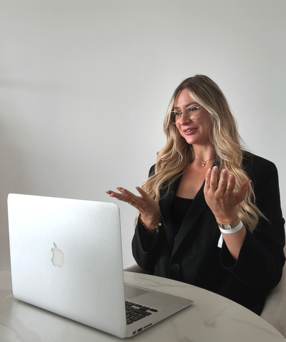 Frau mit brennendem Haar, Brille und Schmuck sitzt vor einem MacBook und gestikuliert bei einer Videokonferenz oder einem Gespräch.