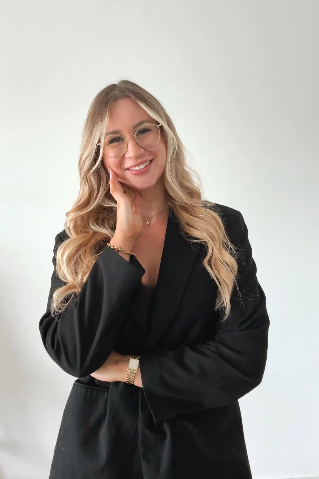 Eine lachende Frau mit langen blonden Haaren, trägt eine schwarze Jacke, Gläser und Schmuck, posiert vor einem weißen Hintergrund.
