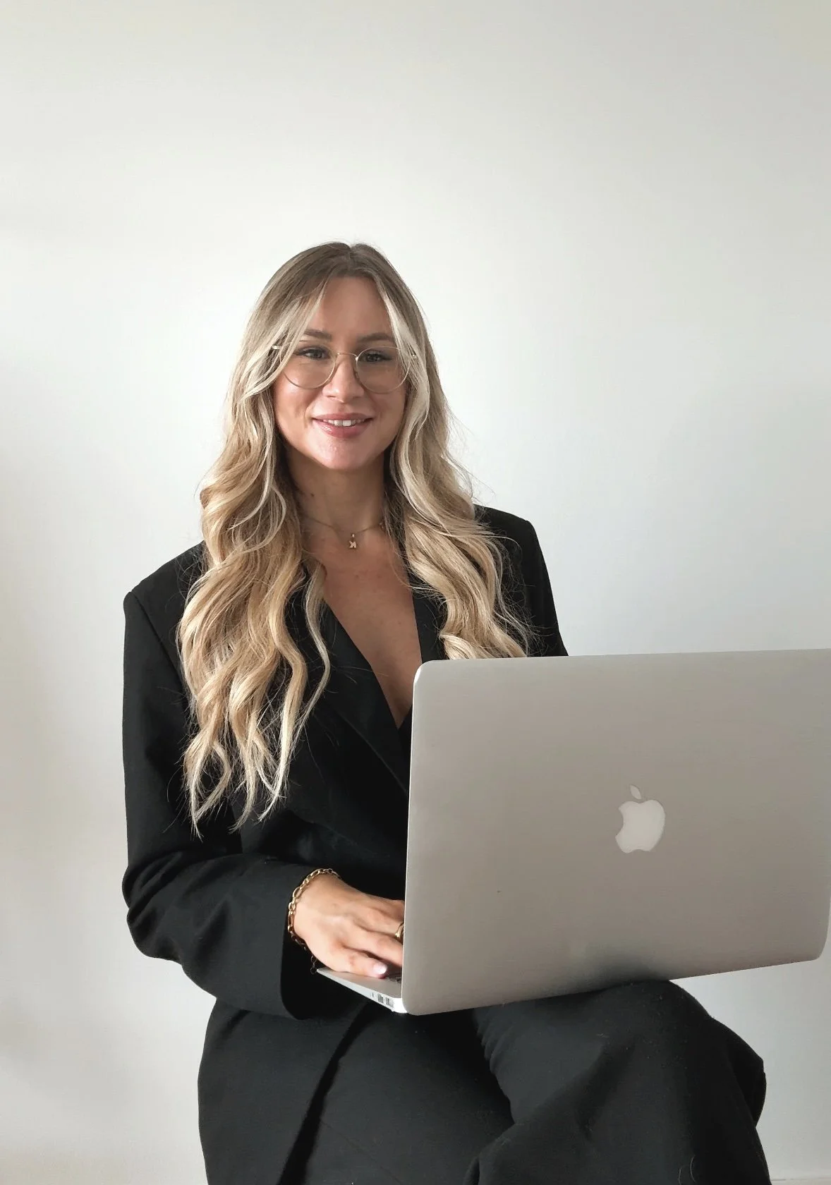 Eine Frau mit langen blonden Haaren und Brille sitzt vor einem Laptop und lächelt. Sie trägt eine schwarze Jacke und einen goldenen Anhänger. Der Hintergrund ist eine weiße Wand.