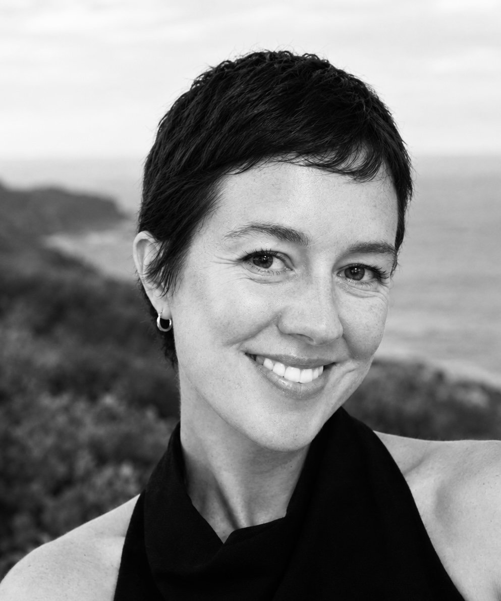 Black and white photo of a woman with short dark hair, smiling, standing outdoors with a blurred landscape background.