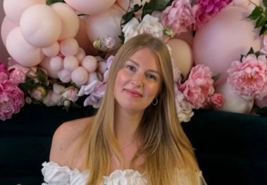 En kvinne med langt blond hår som smiler, foran en blomsterdekorasjon med rosa og hvite blomster.