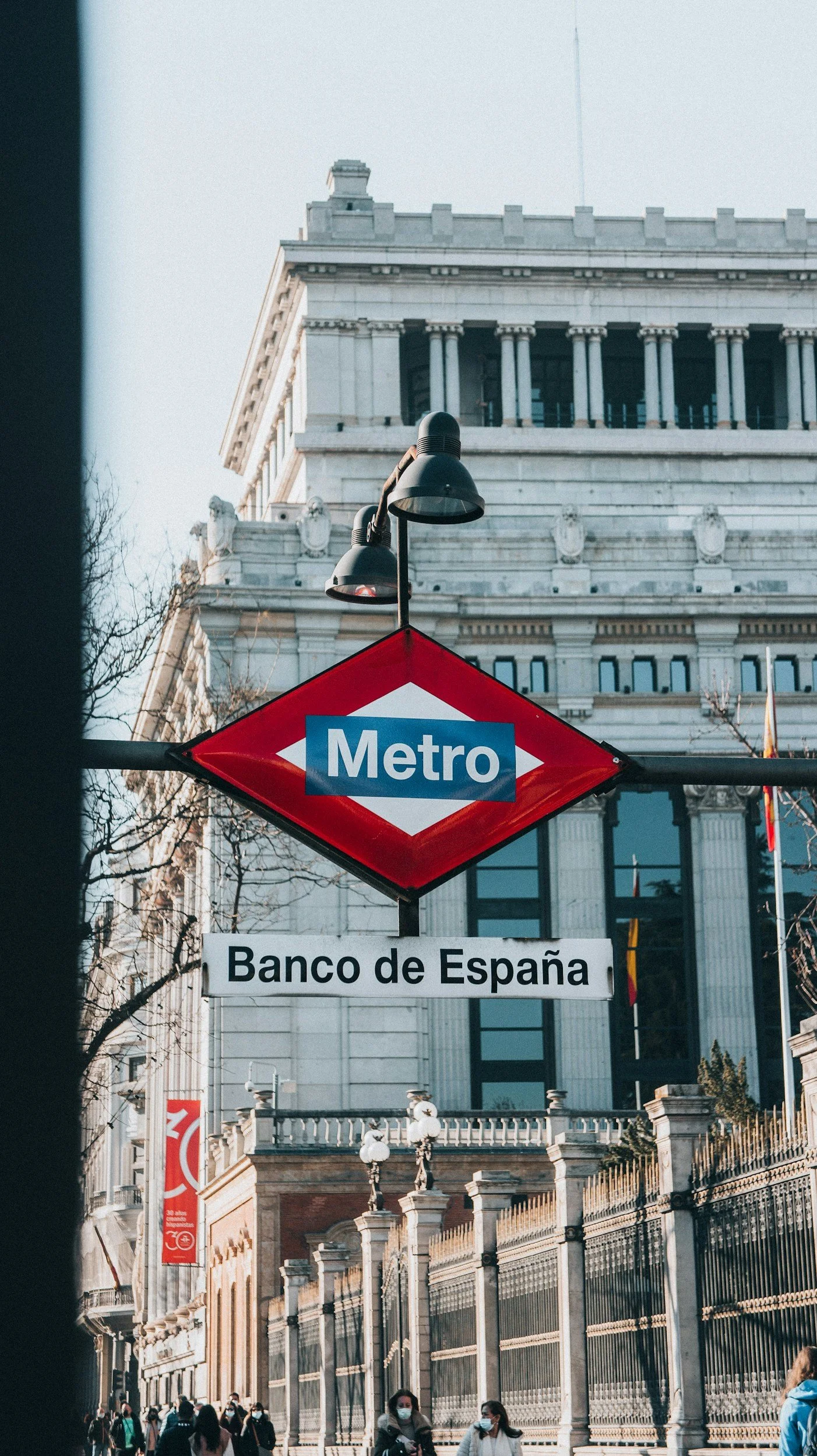 The image shows a metro station sign for Banco de España in Madrid, Spain. The sign is red with a blue and white metro logo. In the background, there is a historic building with classical architecture, featuring columns and decorative stonework. Several people wearing face masks are seen walking near the station.