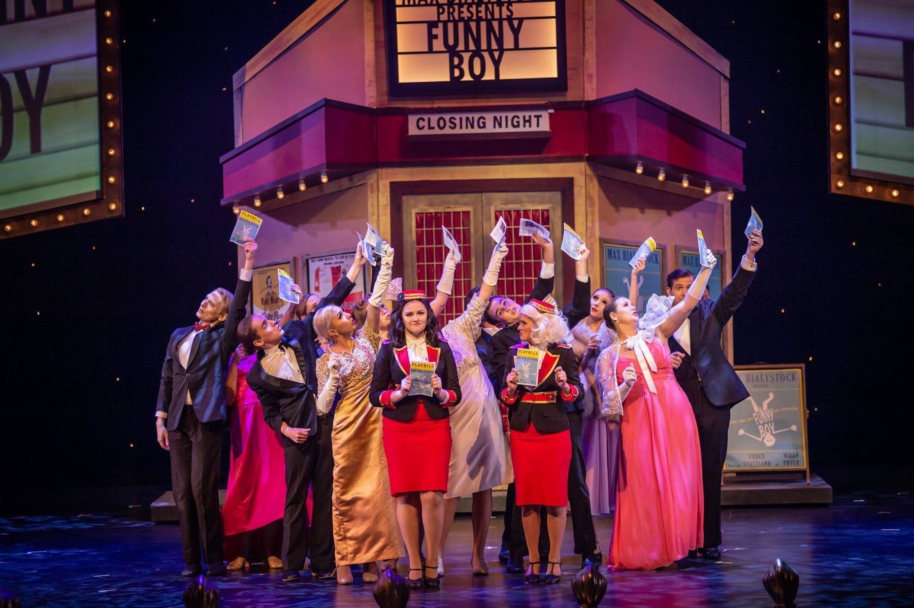 Stage scene from a theatrical production of 'Funny Boy' showing actors in vintage costumes, holding program booklets, with a marquee sign above and a decor backdrop resembling theatre signage.