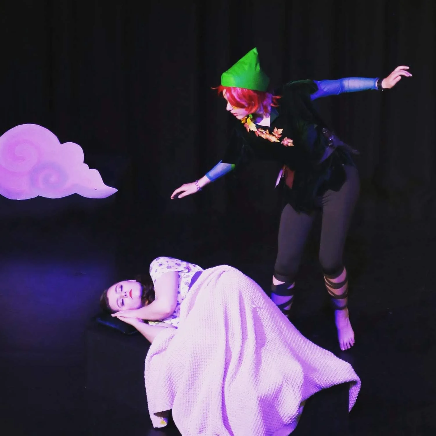 A woman with dark hair is lying on a black floor, covered with a light pink blanket, while a person dressed as a colorful fairy or elf stands nearby with outstretched arms, dressed in a green hat, pink wig, and vibrant costume. The background is dark