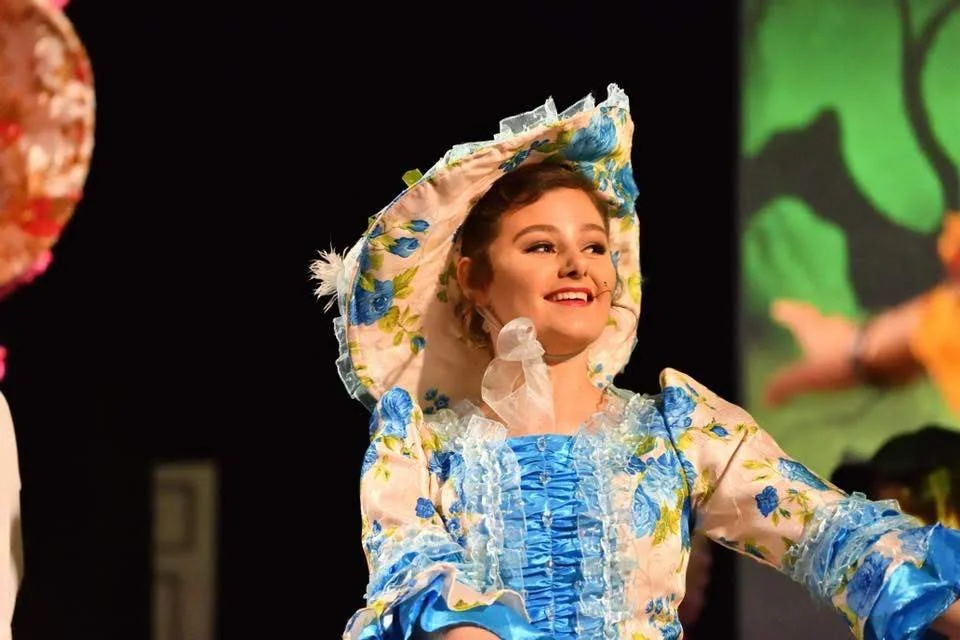 A young woman on stage, smiling, wearing a floral Victorian-style dress with a bonnet, during a theatrical performance.