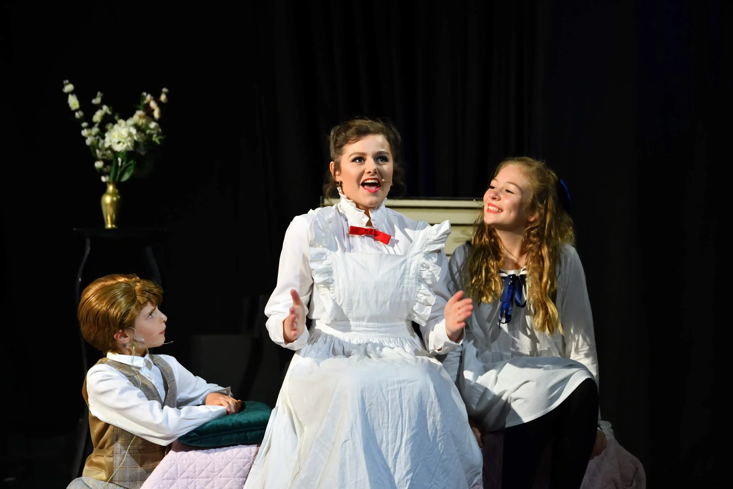 Scene from a theatrical play featuring four young girls in vintage costumes, with one girl standing and speaking, two girls seated, and a fourth partially visible at the bottom. A vase with flowers is on a table to the side, set against a dark backgr