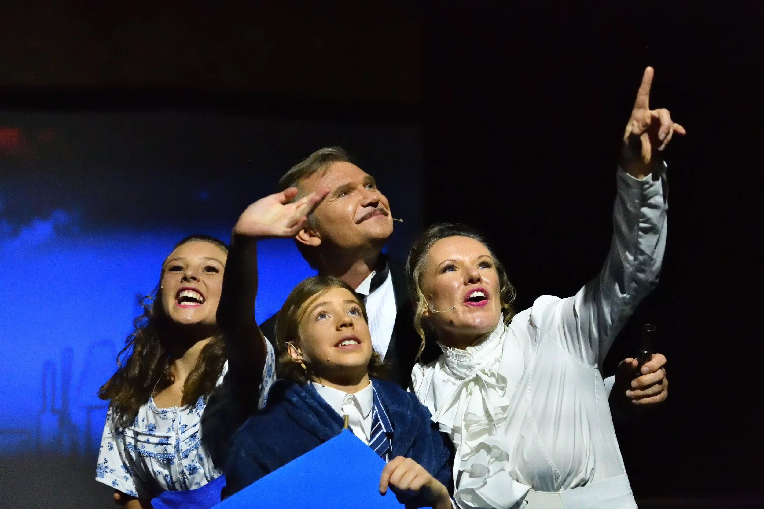 A group of people, including adults and children, on stage. They are dressed in formal or semi-formal attire, with a dark background and stage lighting.