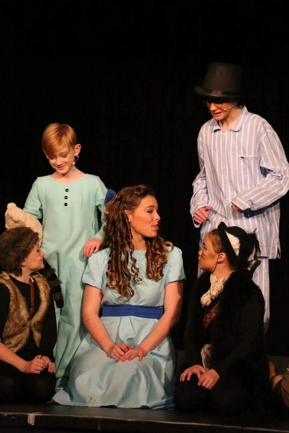 Children and an adult in costume on stage during a theatrical performance, with the children dressed as characters and the adult portraying a character in striped pajamas and a hat, engaged in a scene.