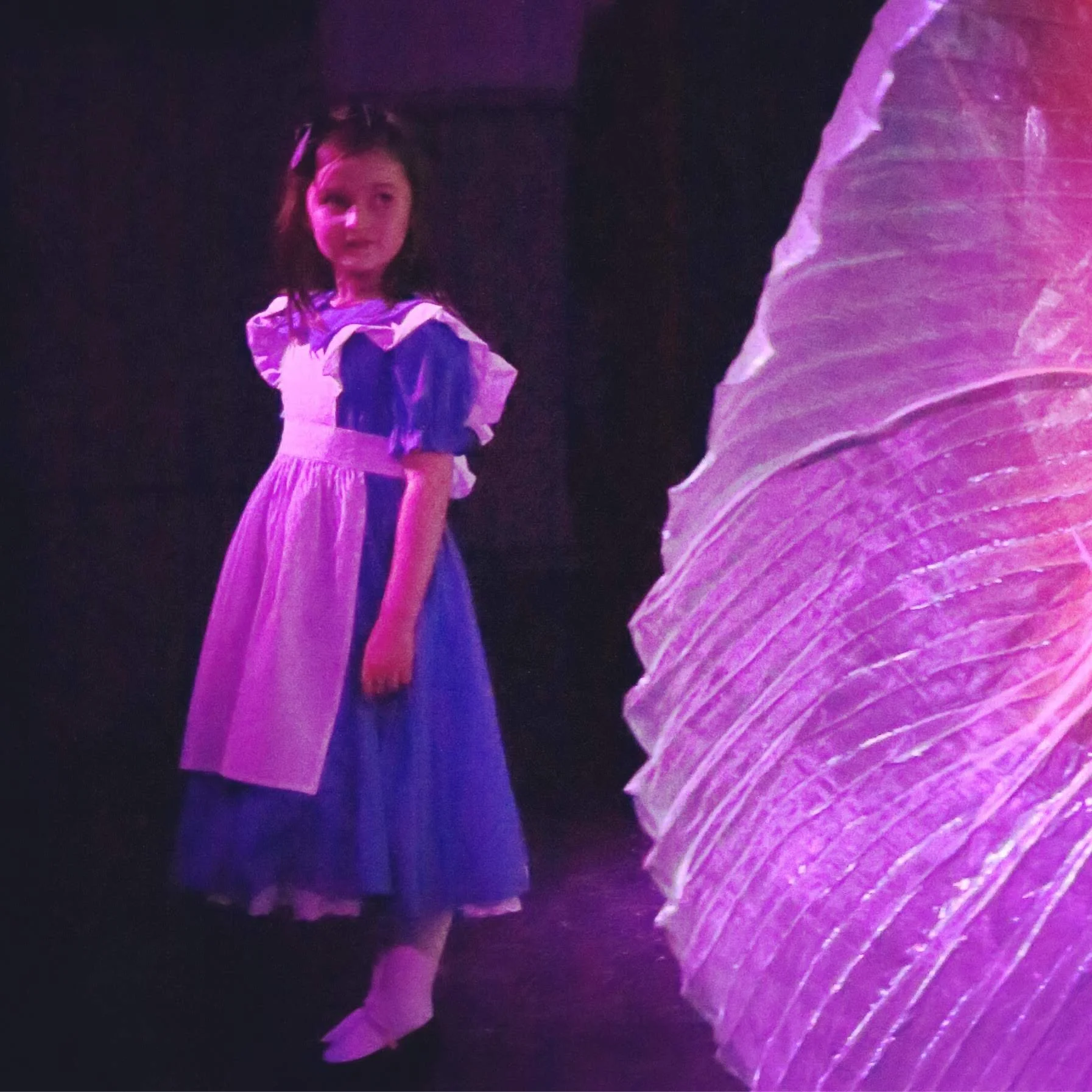 A young girl with dark hair and a dress with puffed sleeves, standing in a dark, purple-lit environment with a large, textured purple surface nearby.