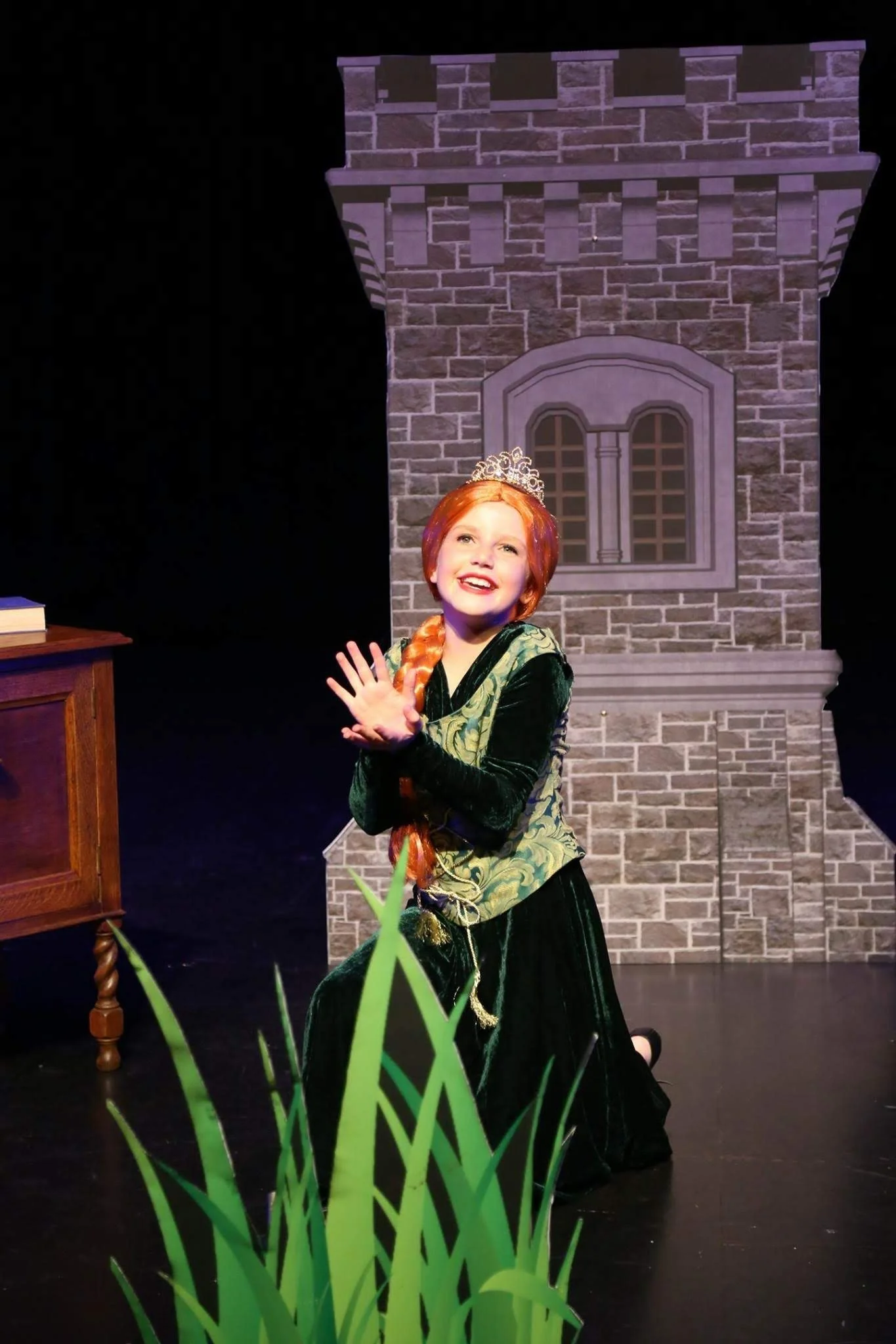 A woman dressed as a princess with red hair, a tiara, and a long black and green dress, posing in front of a brick castle backdrop on a stage.