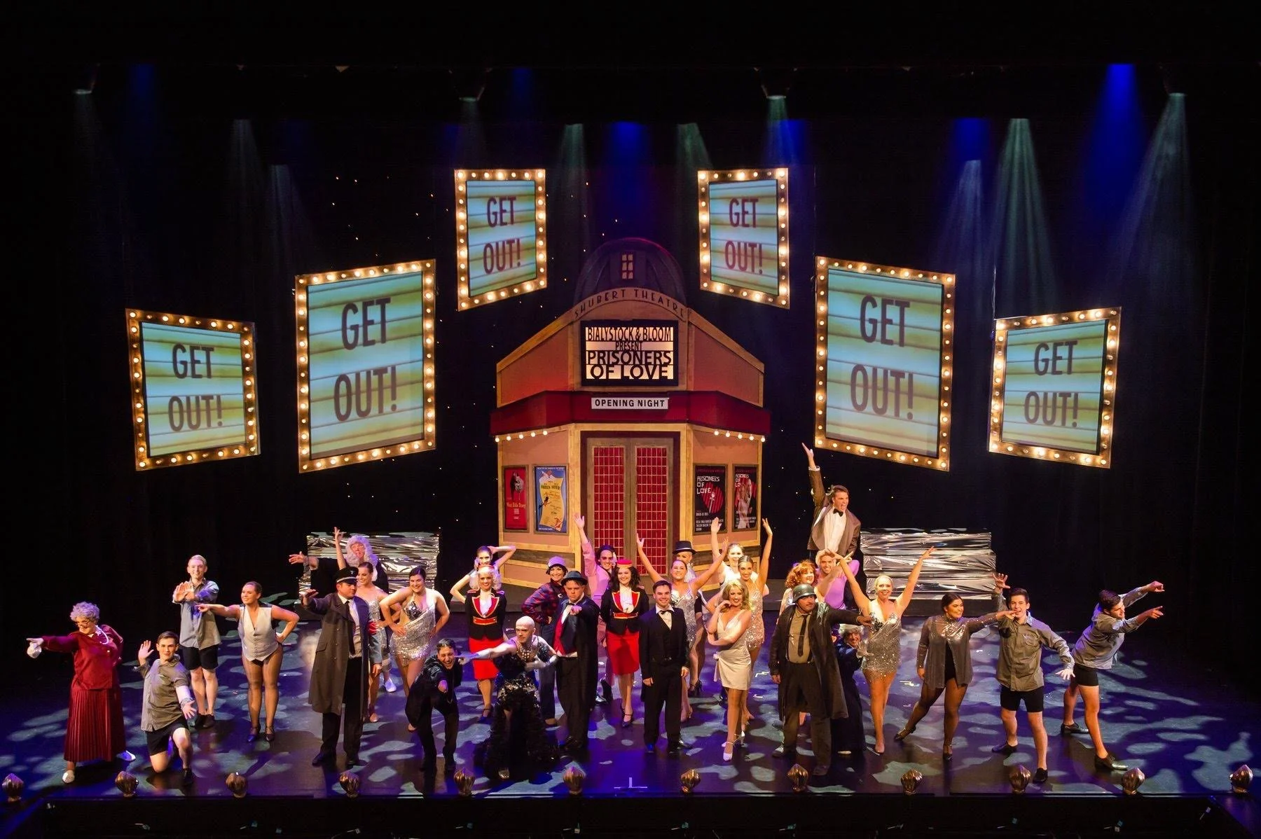Theatre stage with cast members in costume performing; background features illuminated signs reading 'GET OUT!' and a marquee with the show title 'Prisoners of Love' and opening night information.